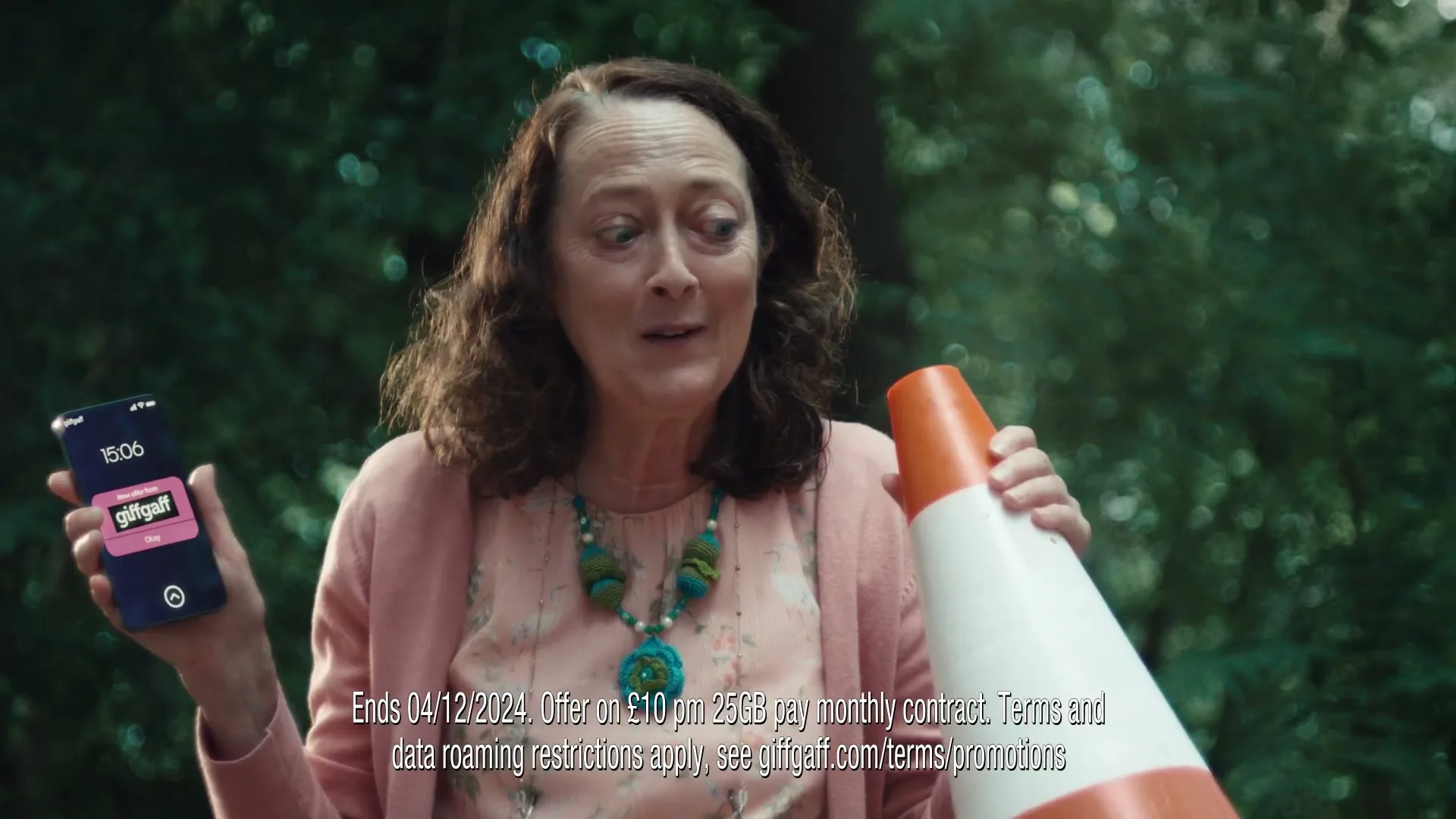 A middle-aged woman in a pink cardigan holds a phone and a traffic cone, looking distressed in a park.