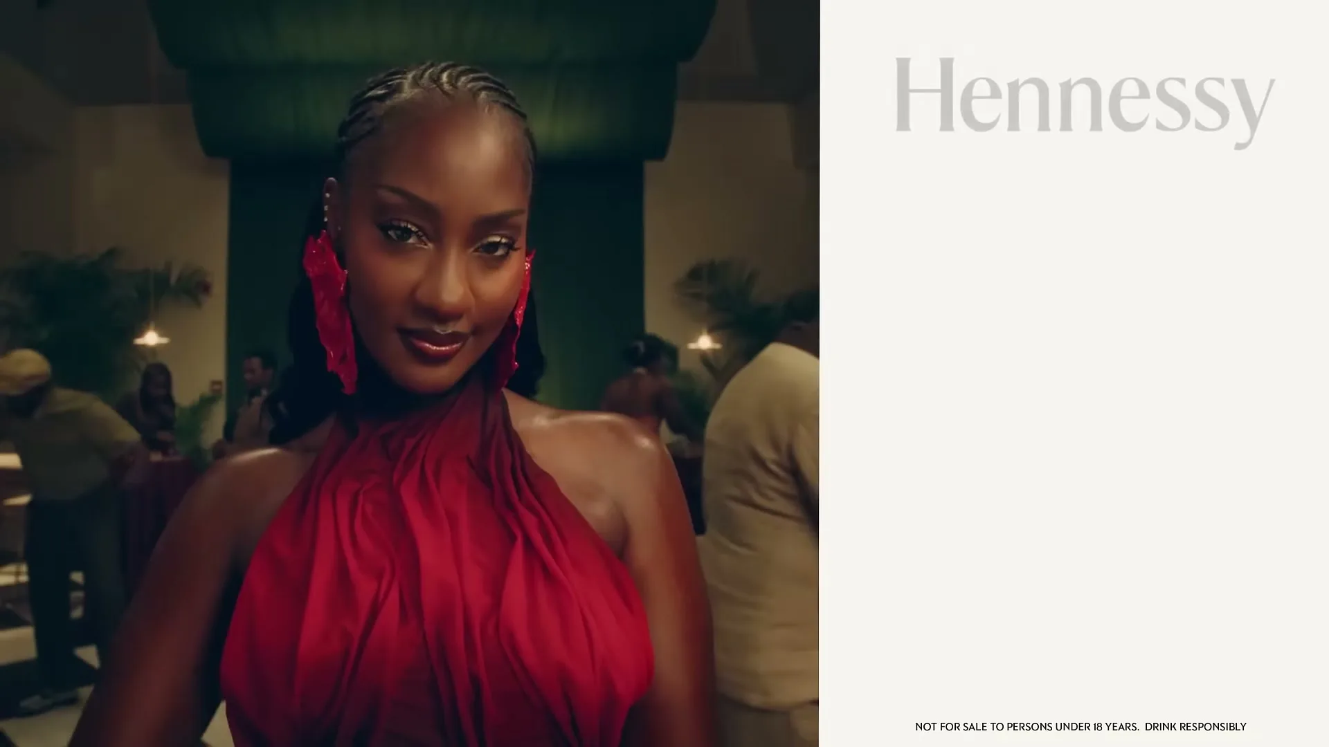 A beautiful Black woman in a red dress and large red earrings smiles at the camera in a dimly lit, elegant interior setting.