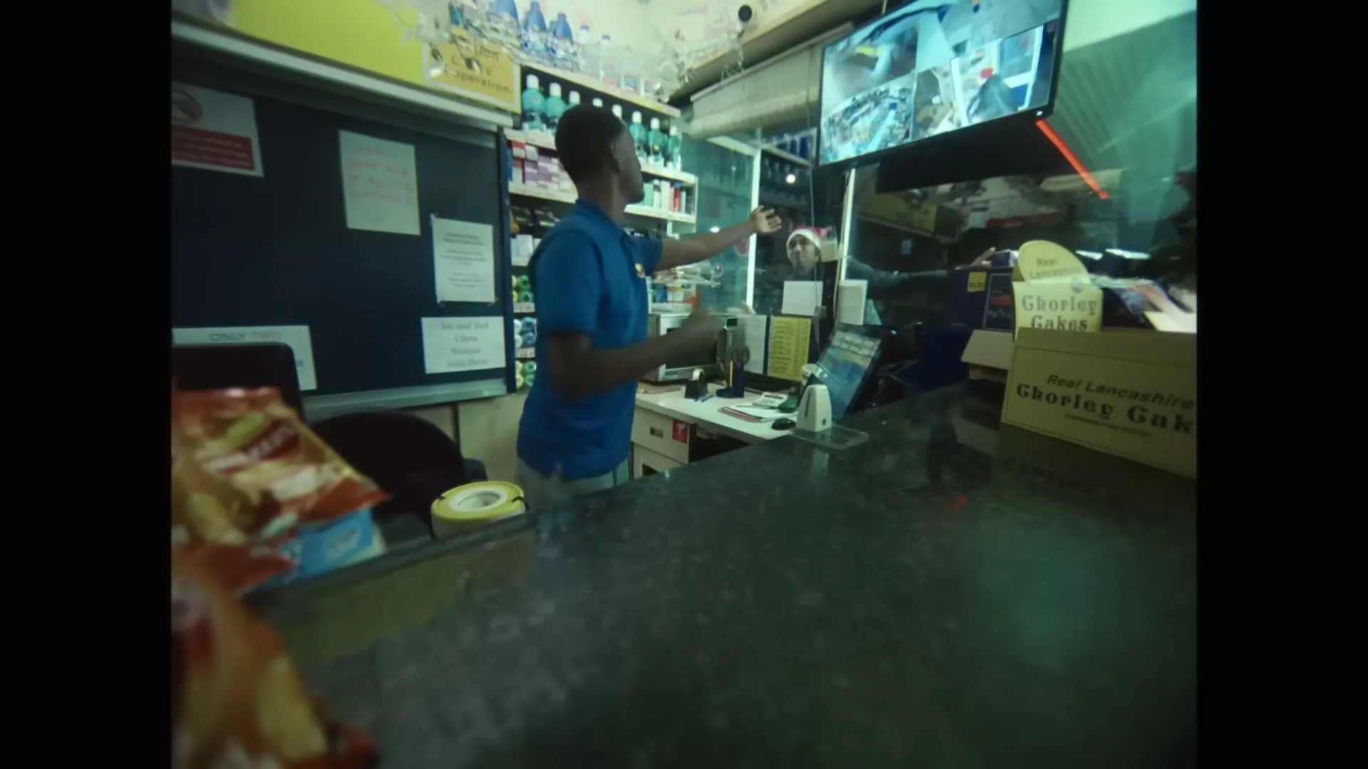A young man in a blue polo shirt stands behind a counter in a convenience store, gesturing towards the entrance as another person enters.