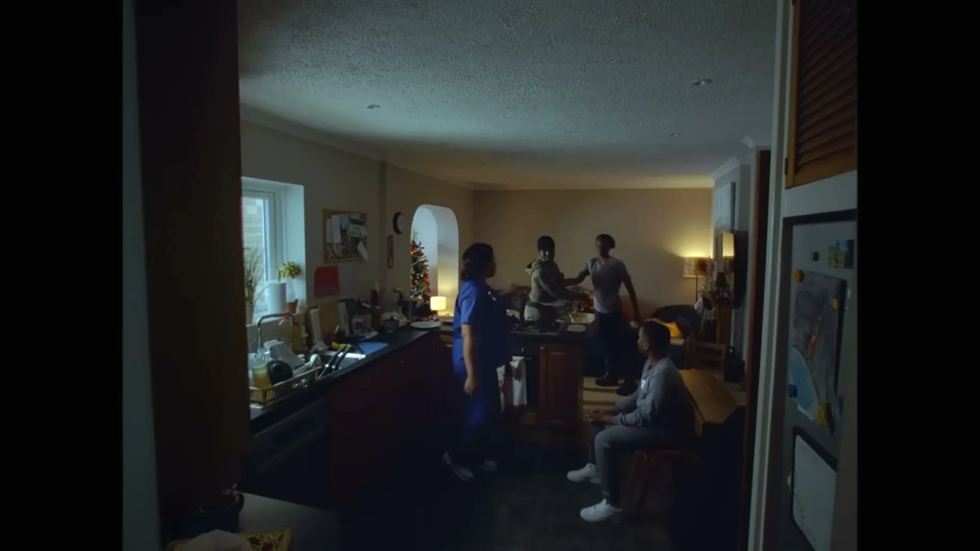 A family of four, dressed in casual clothes, is seen in a dimly lit kitchen and living room, with one person washing dishes and others interacting.