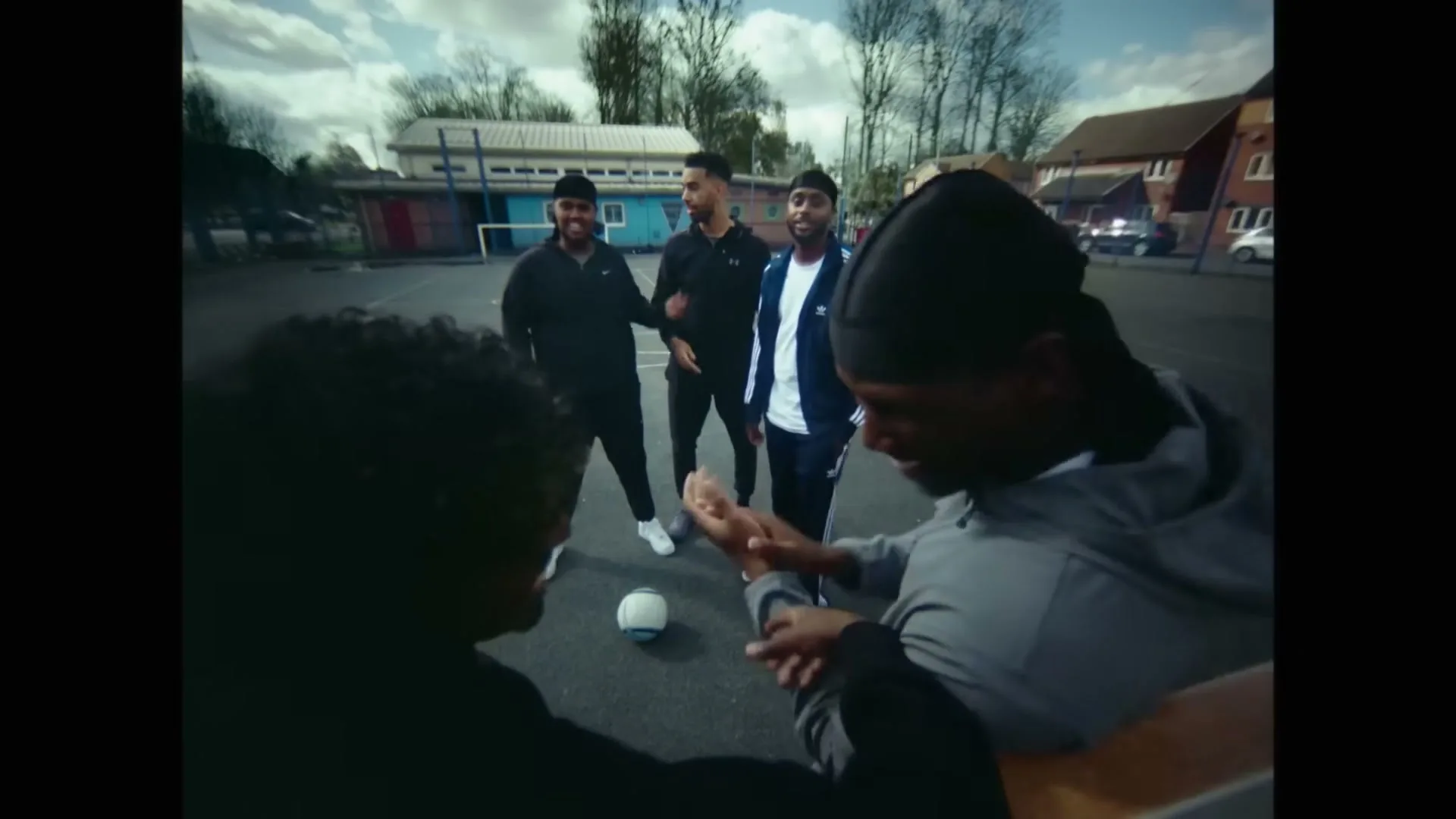 Five young men play football on a concrete court, with two in the foreground and three in the background, all smiling and interacting.