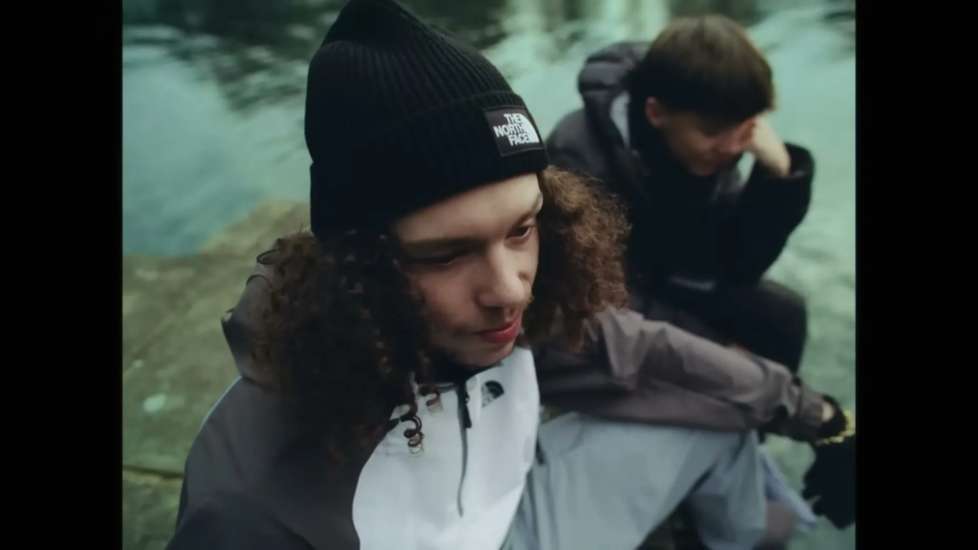 Two young men, one with curly hair and a beanie, sit by a body of water, looking down thoughtfully in an outdoor setting.
