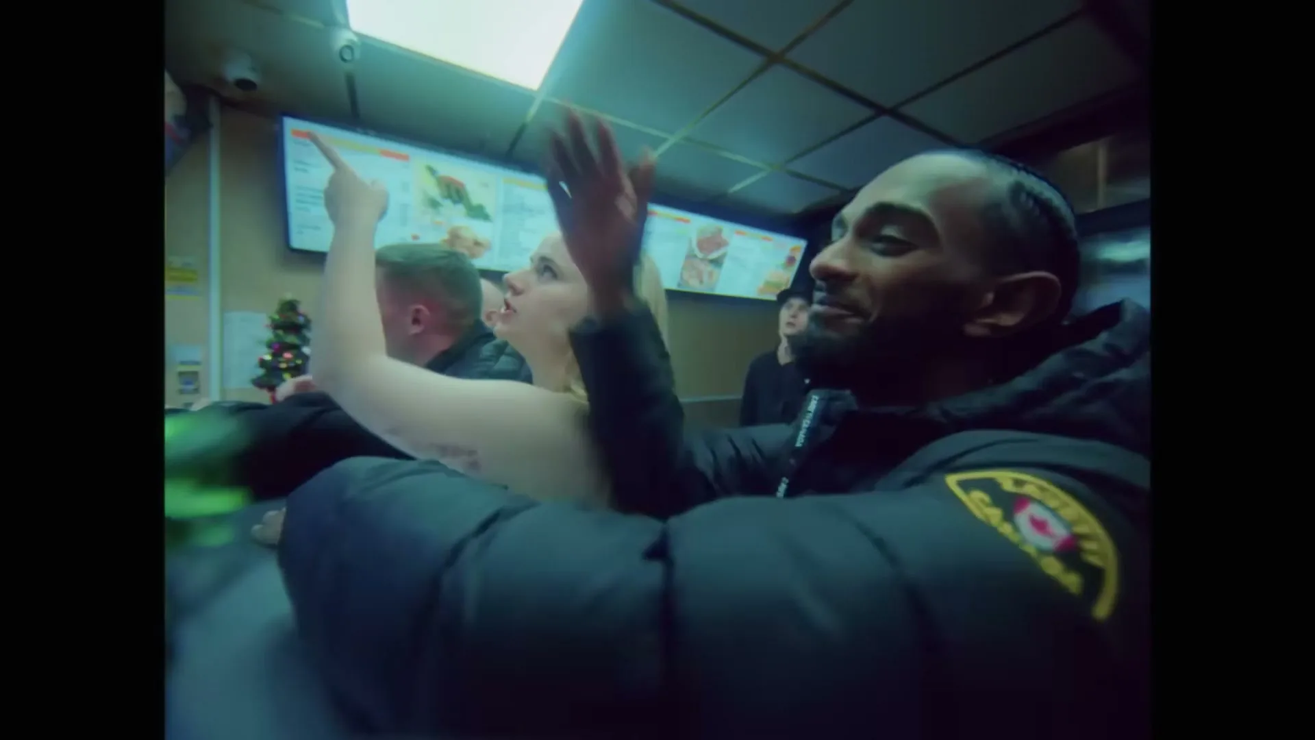 A diverse group of young adults, including a man in a black puffer jacket and a woman with blonde hair, are excitedly pointing and gesturing inside a brightly lit fast-food restaurant.