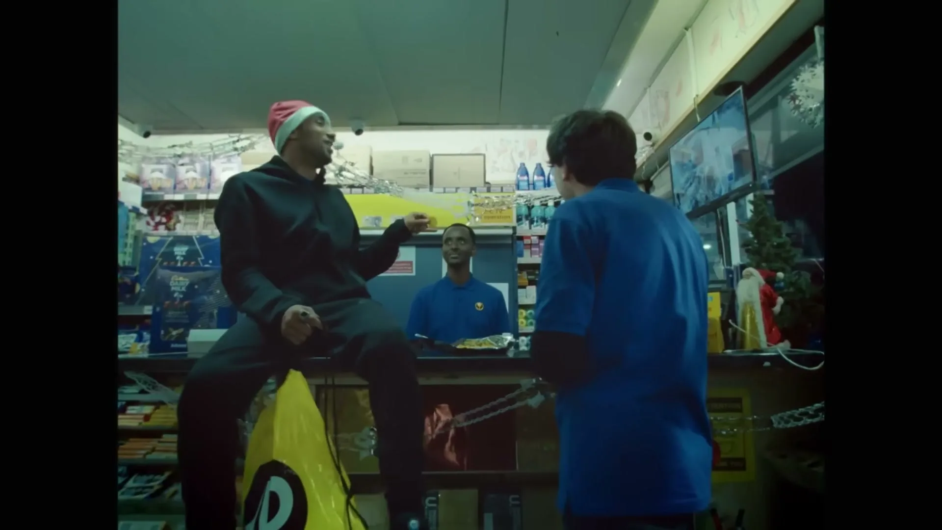 A man in a Santa hat sits on a yellow cone, talking to another man in a blue shirt, while a third man stands behind a counter in a convenience store.
