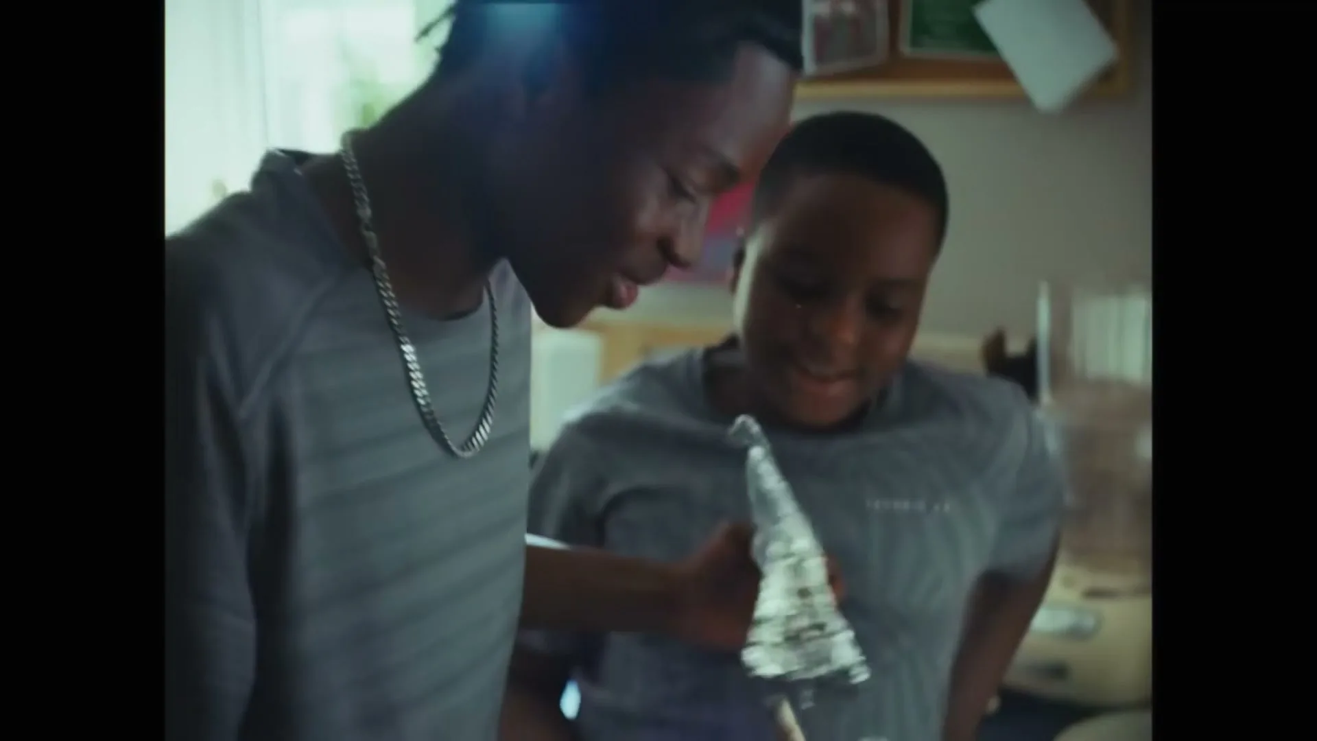Two young Black males, one older with dreadlocks and a silver chain, the other younger, are in a kitchen, cooking and playing with whipped cream.