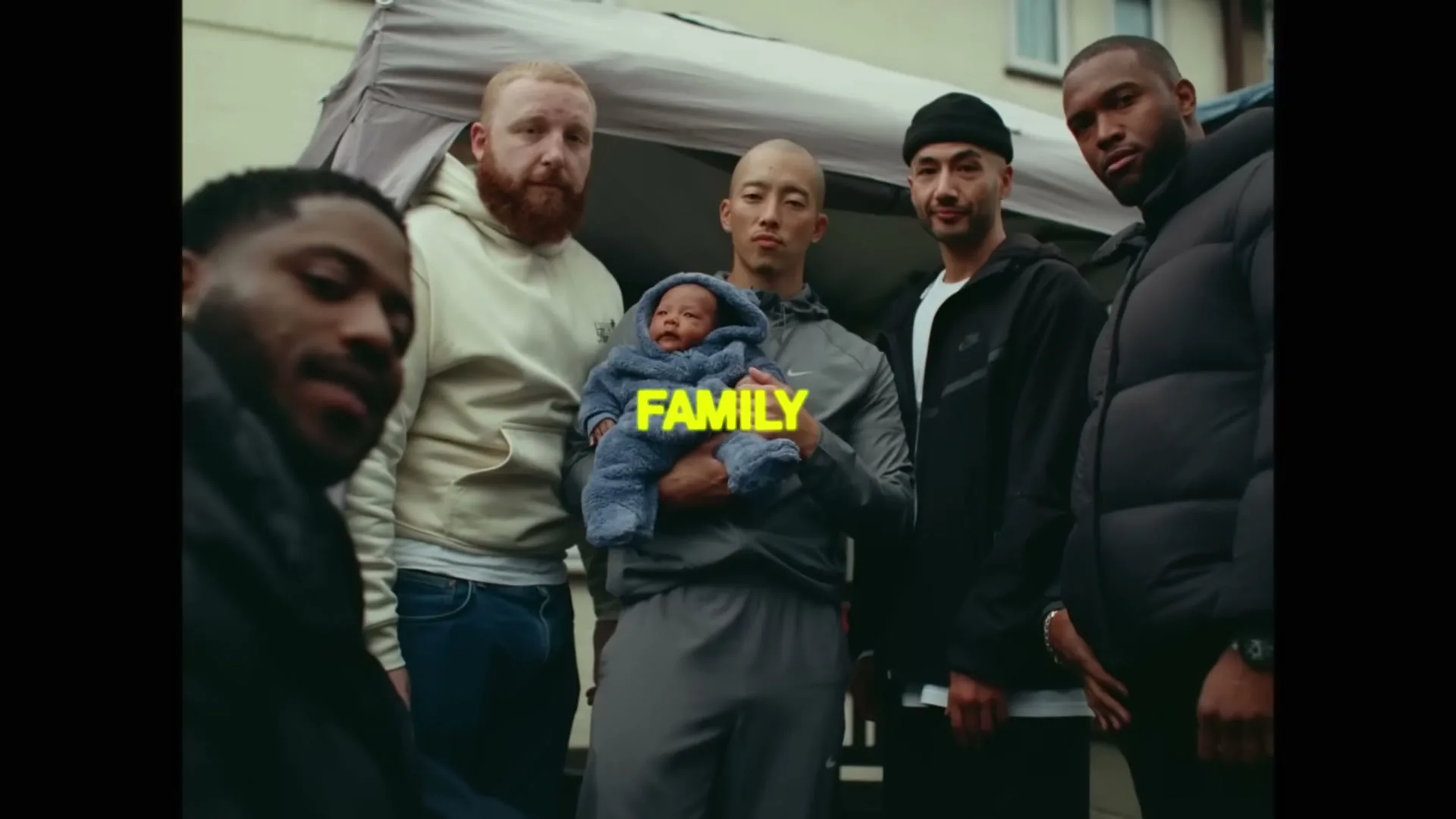 Five men and a baby stand outside a house, looking directly at the camera, with the word "FAMILY" superimposed in yellow.