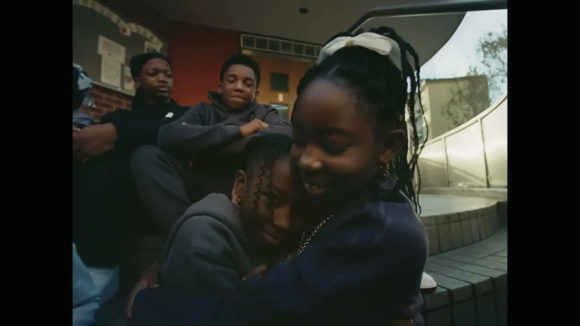 Two young Black girls hug each other, smiling, while other young people sit on steps in the background, outside a building.