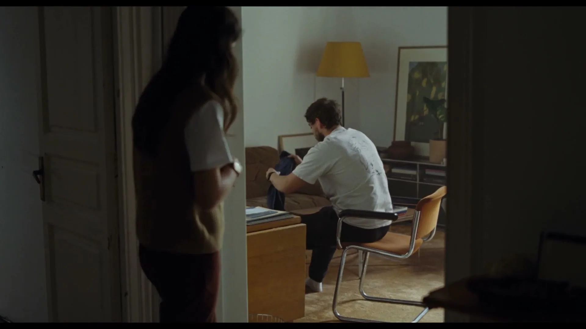 A woman stands in a doorway, looking into a room where a man sits at a desk, packing a bag.