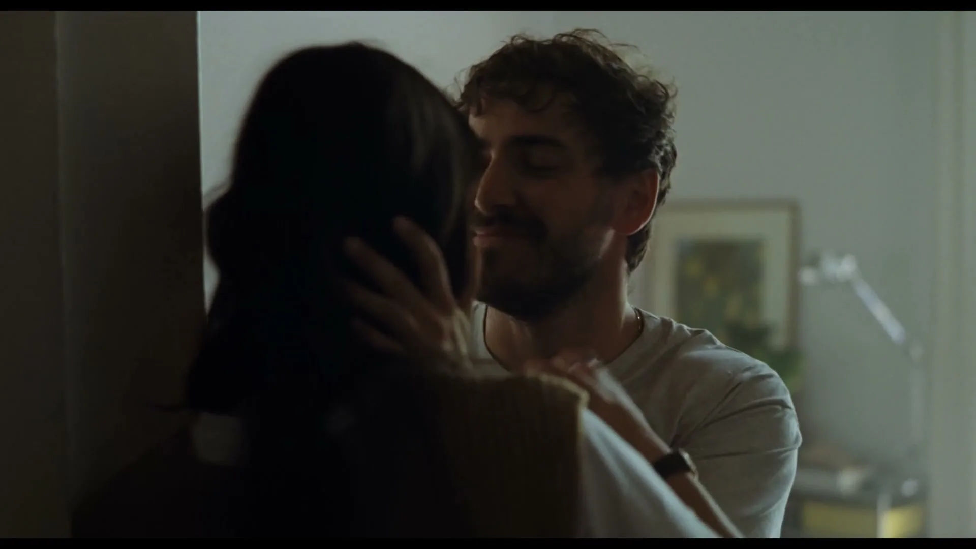 A man with a beard and curly hair embraces a woman with long dark hair, kissing her cheek in a warmly lit interior.
