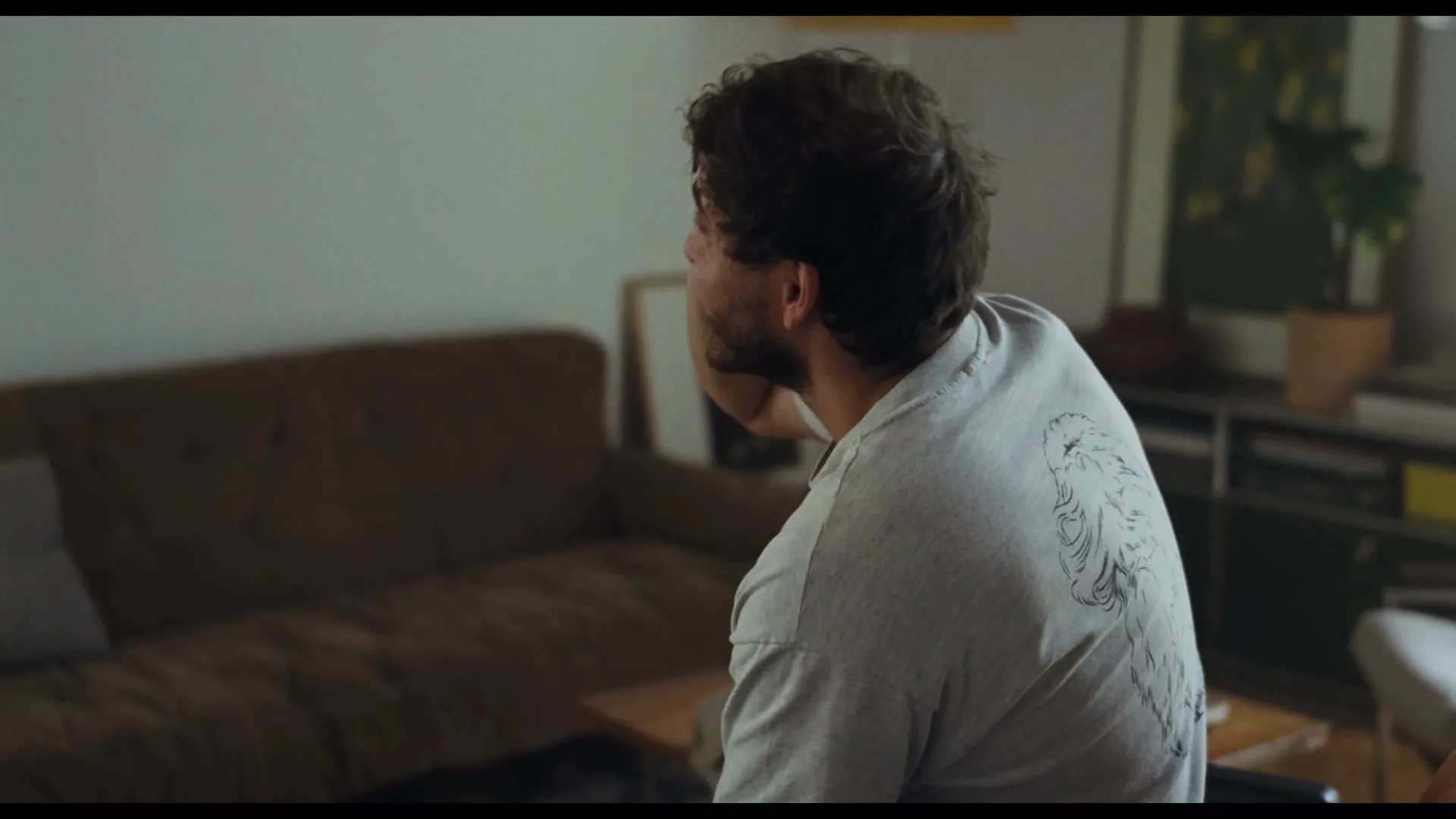 A man with dark, curly hair and a beard, wearing a light grey t-shirt, sits with his back to the camera, looking up and to the left in a living room.