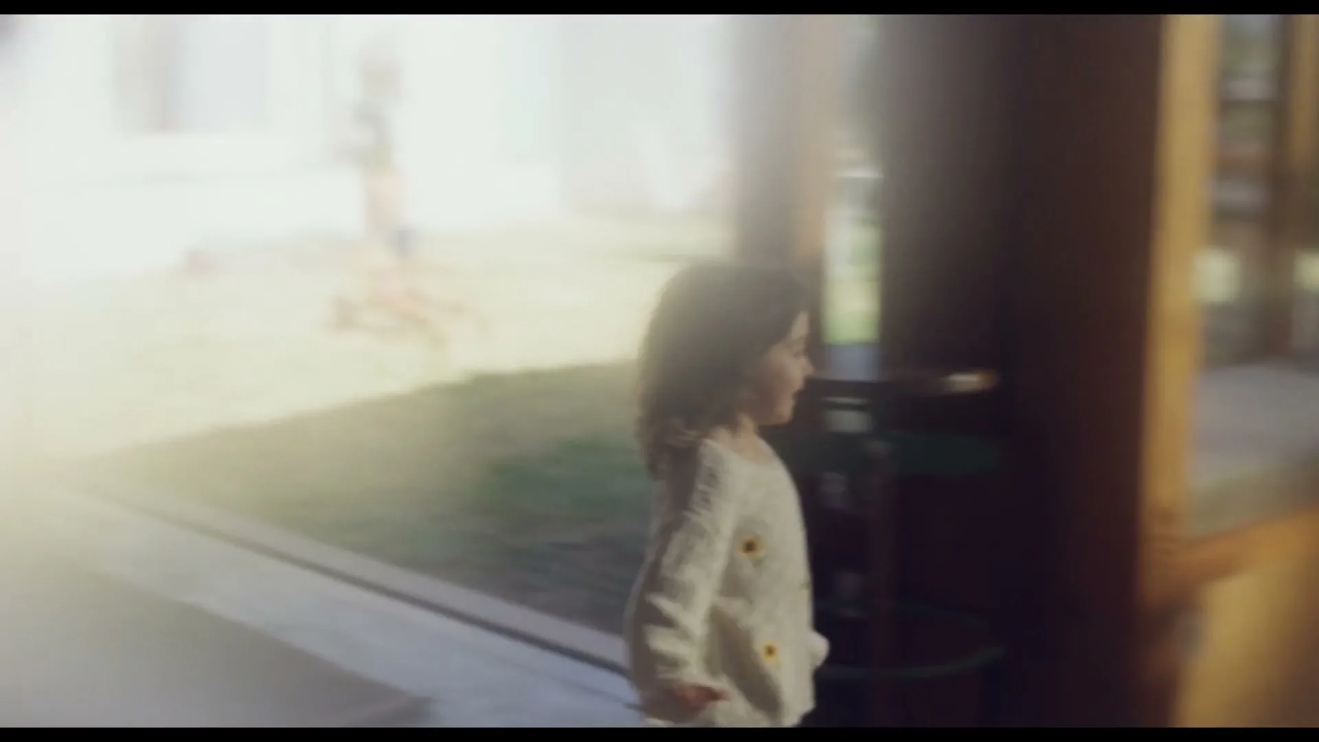 A young child with curly brown hair and a cream sweater walks past a large window, looking out at a sunny backyard.
