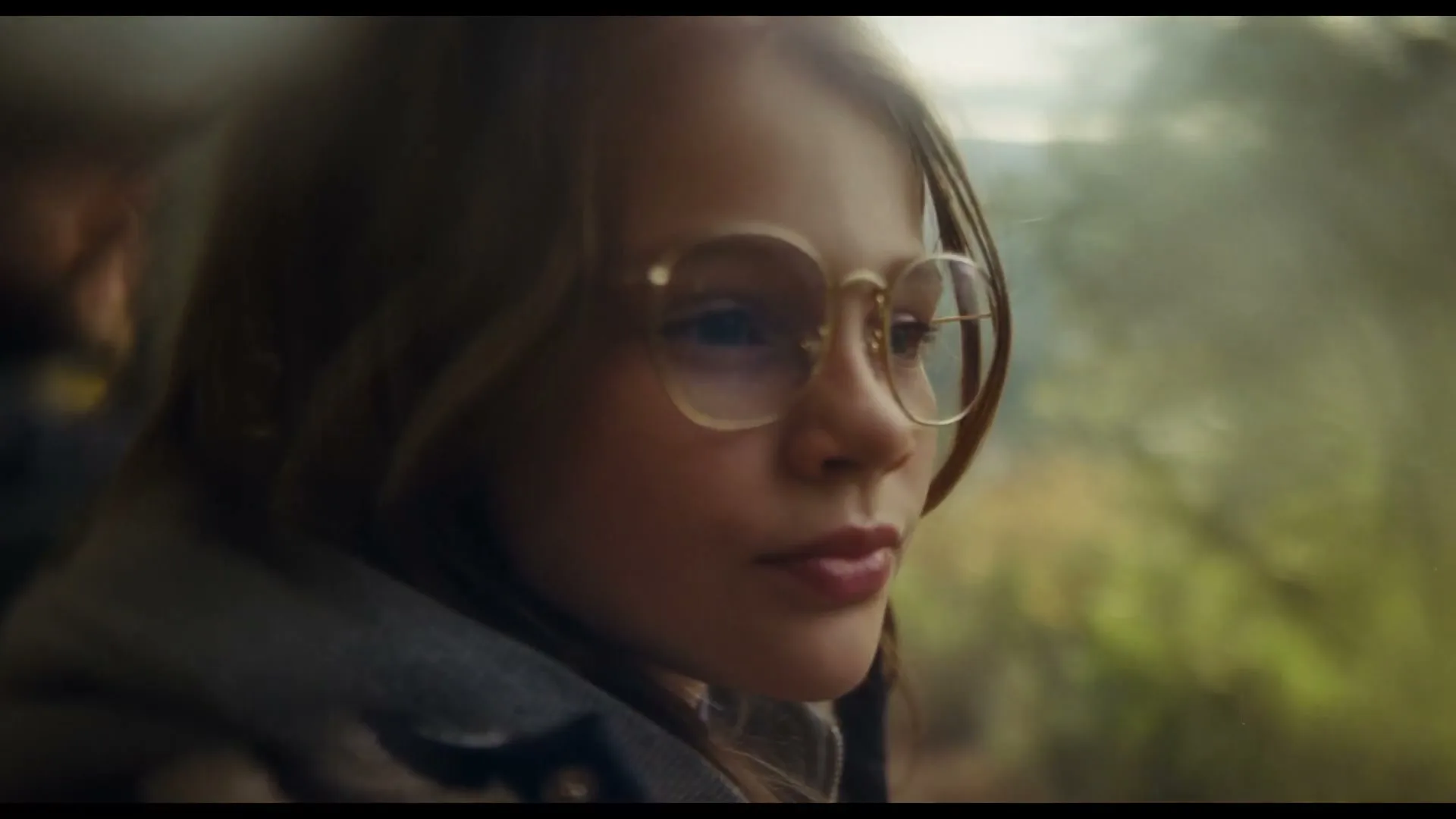 A young girl with long hair and glasses looks out a window, with sunlight and lens flares creating a dreamy effect.