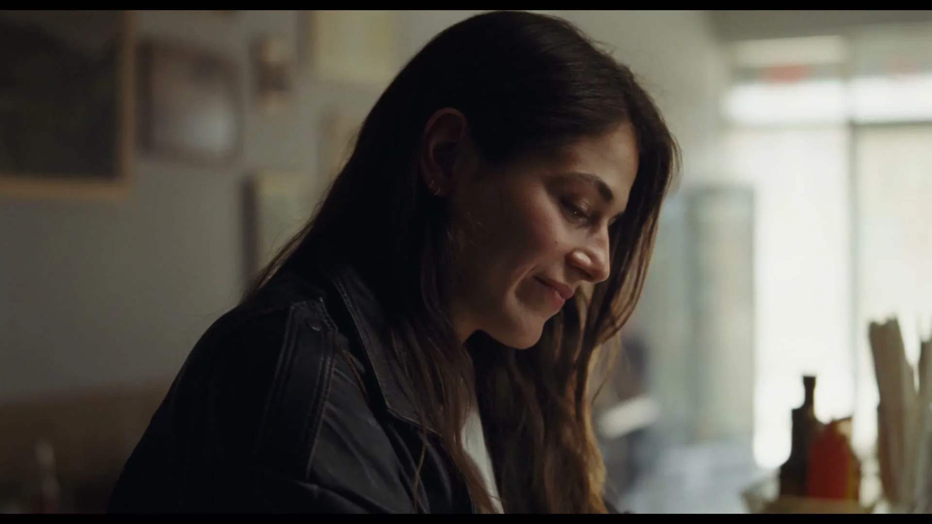 A young woman with long dark hair, wearing a dark jacket, smiles softly while looking down, in a dimly lit interior space.