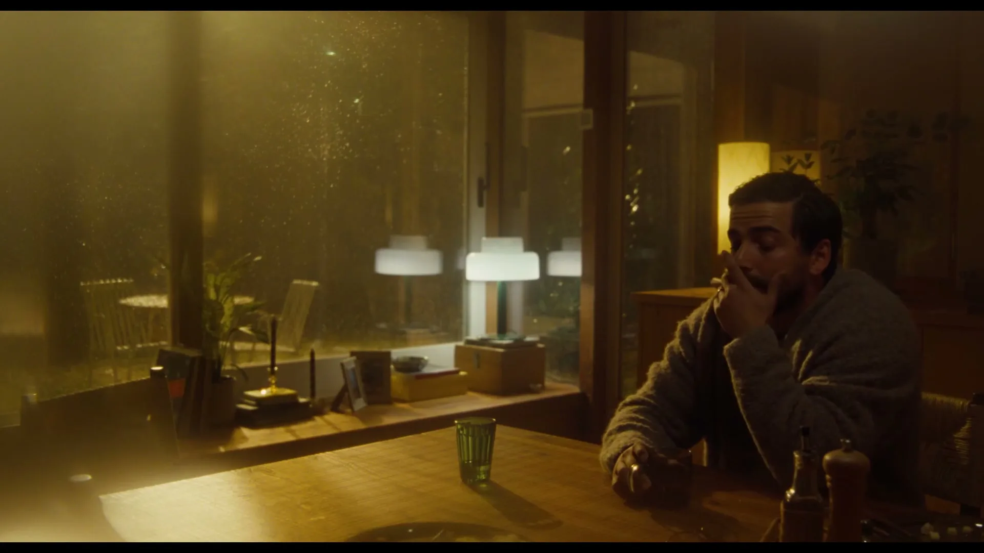 A man with dark hair and a beard sits at a wooden table, looking pensive, in a dimly lit room with large windows.
