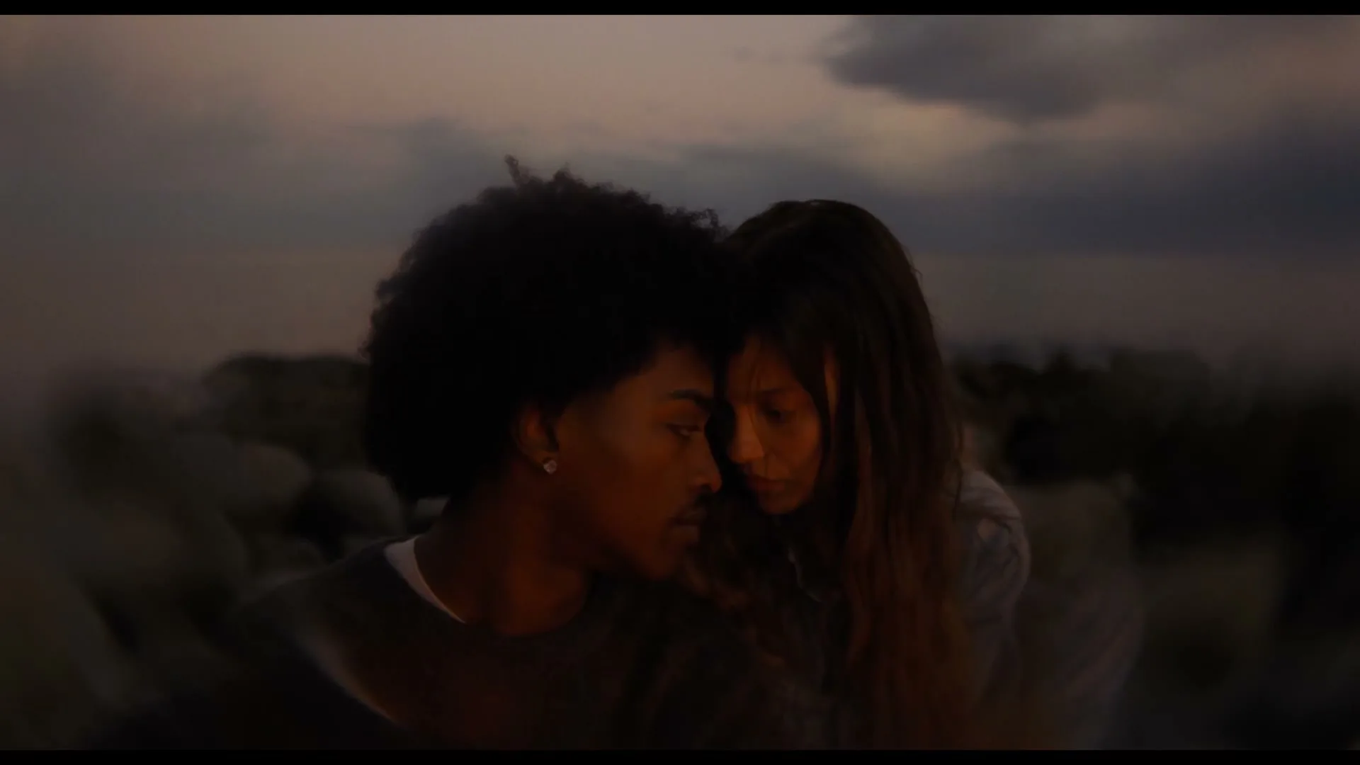 A young couple, a man with an afro and a woman with long hair, share an intimate moment, foreheads touching, on a rocky beach at dusk.