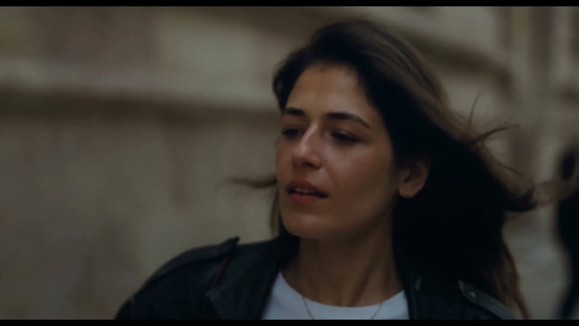 A woman with dark hair and a black leather jacket looks around with a concerned expression on a city street.