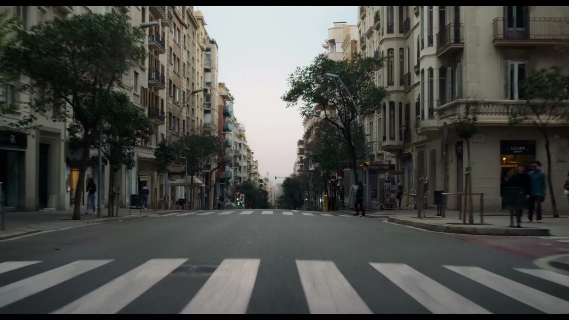A wide shot of a quiet city street with buildings lining both sides, a crosswalk in the foreground, and a few pedestrians walking.