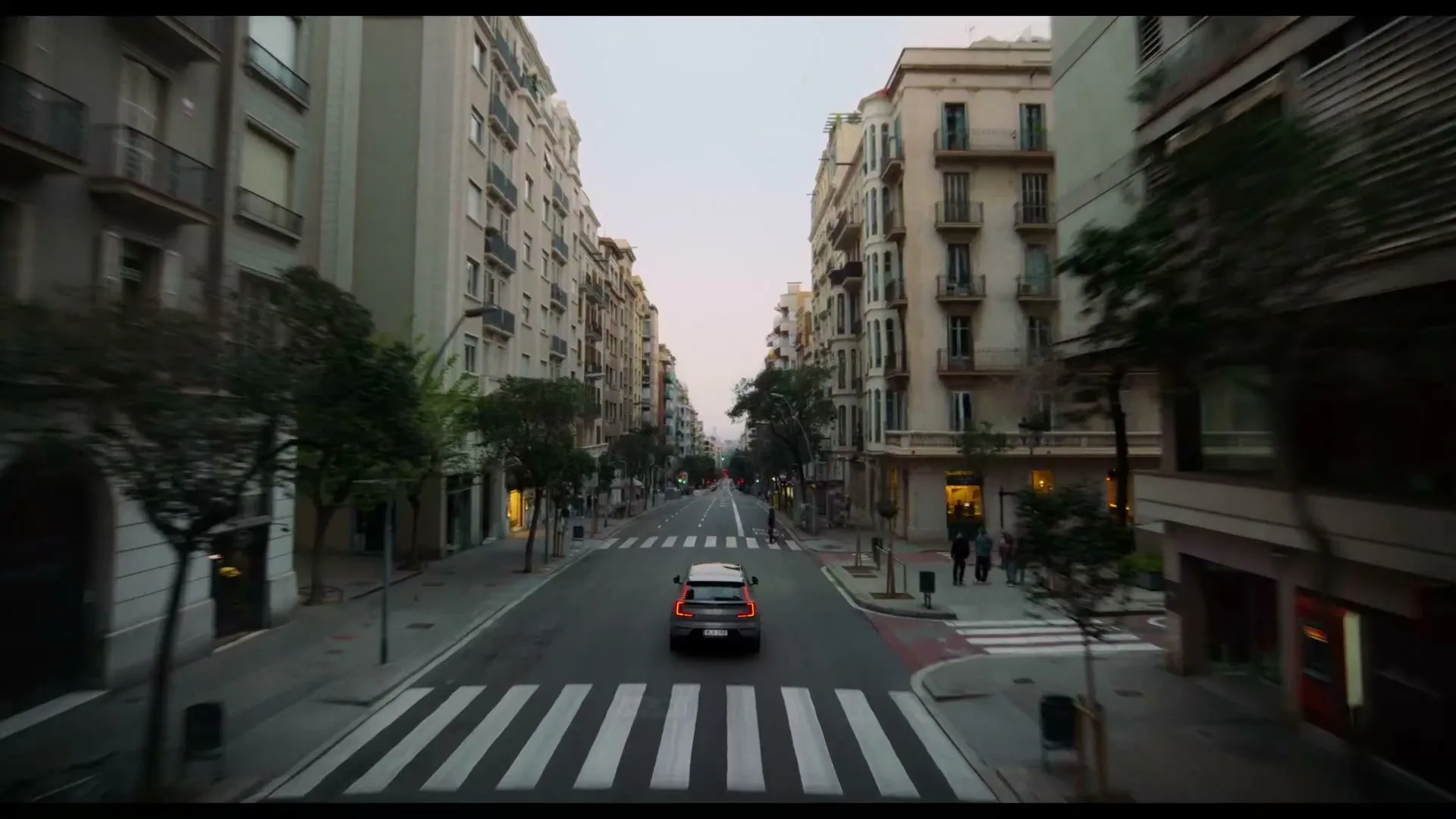 A grey Volvo car drives away from the camera down a wide street lined with tall apartment buildings in a city at dusk.