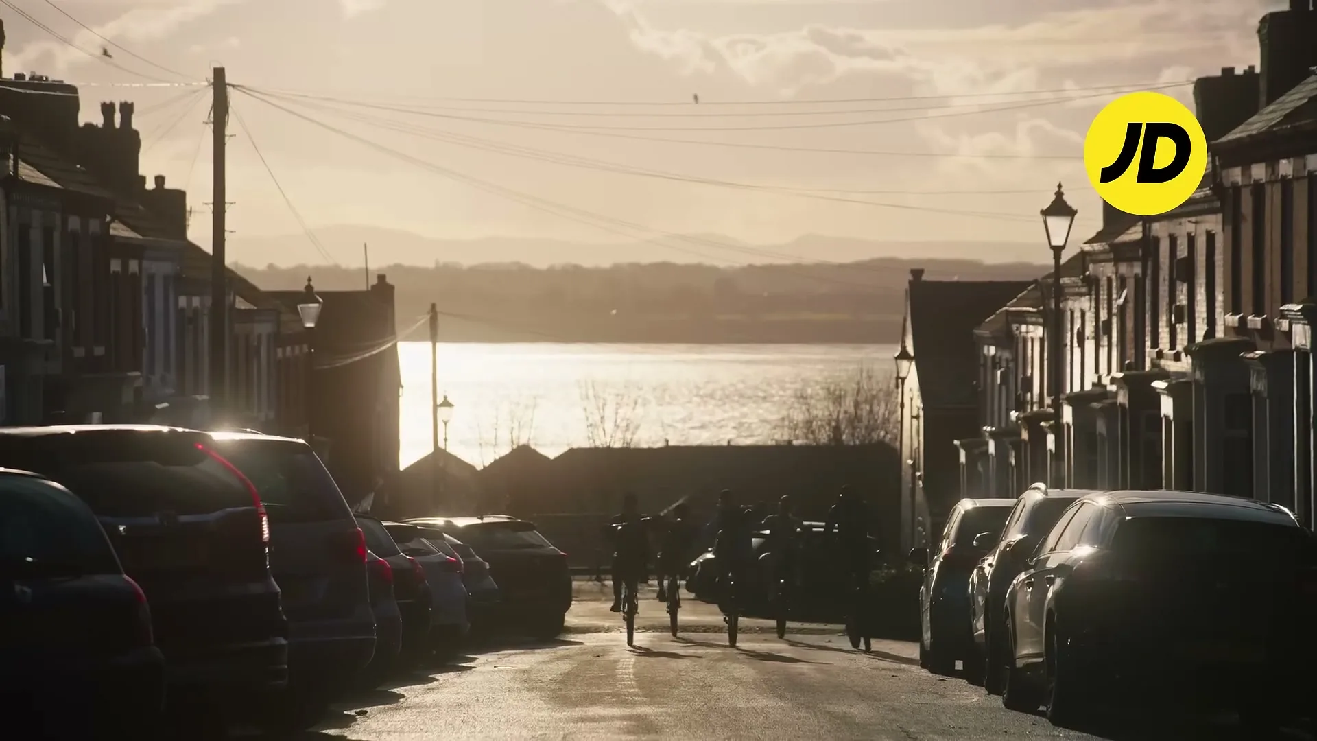 A wide shot of a street with parked cars on both sides, leading down to a body of water, with several people on bicycles riding down the street.