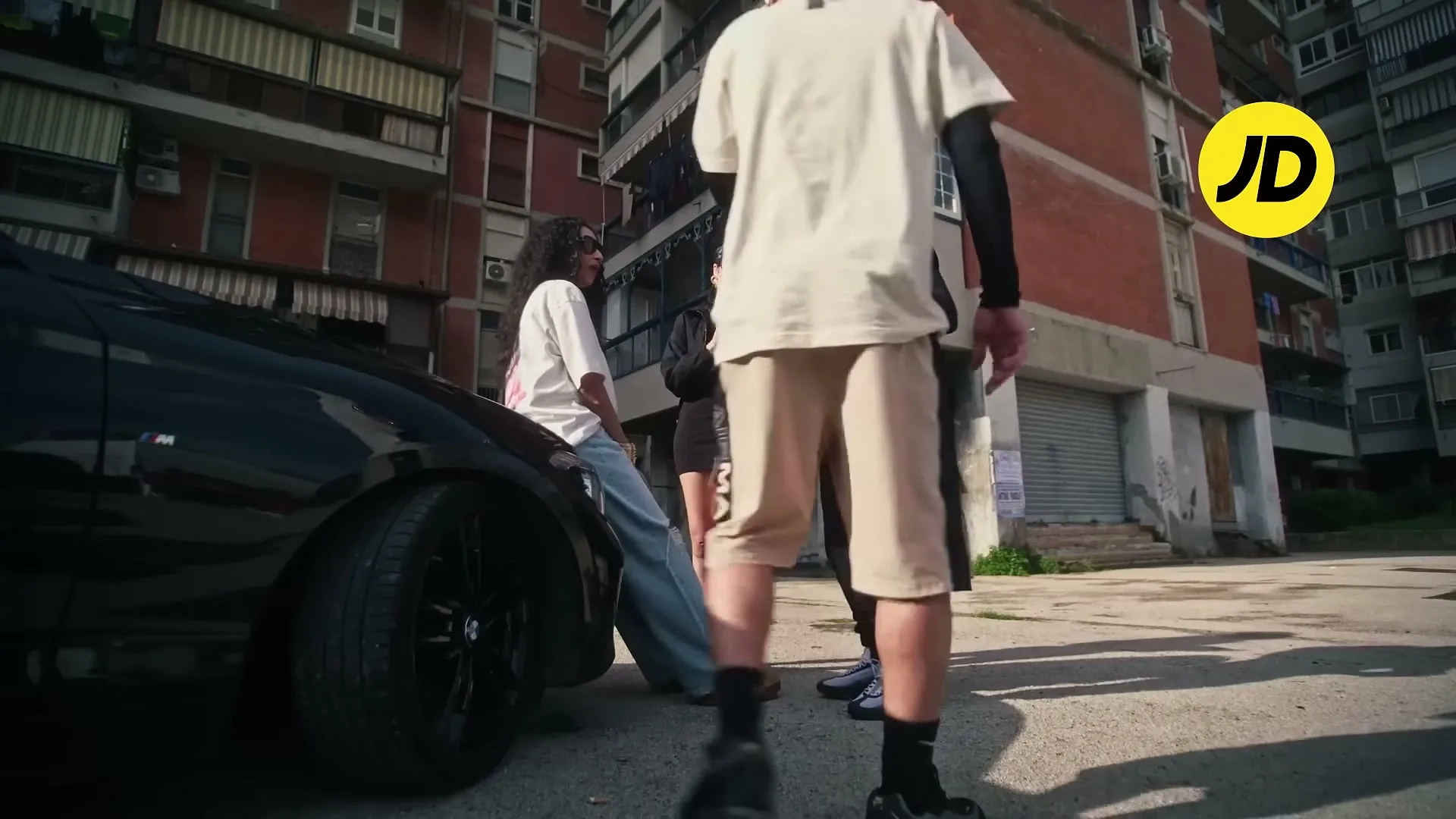 Three young adults in streetwear gather around a black car on a sunny day in a gritty urban setting with apartment buildings in the background.