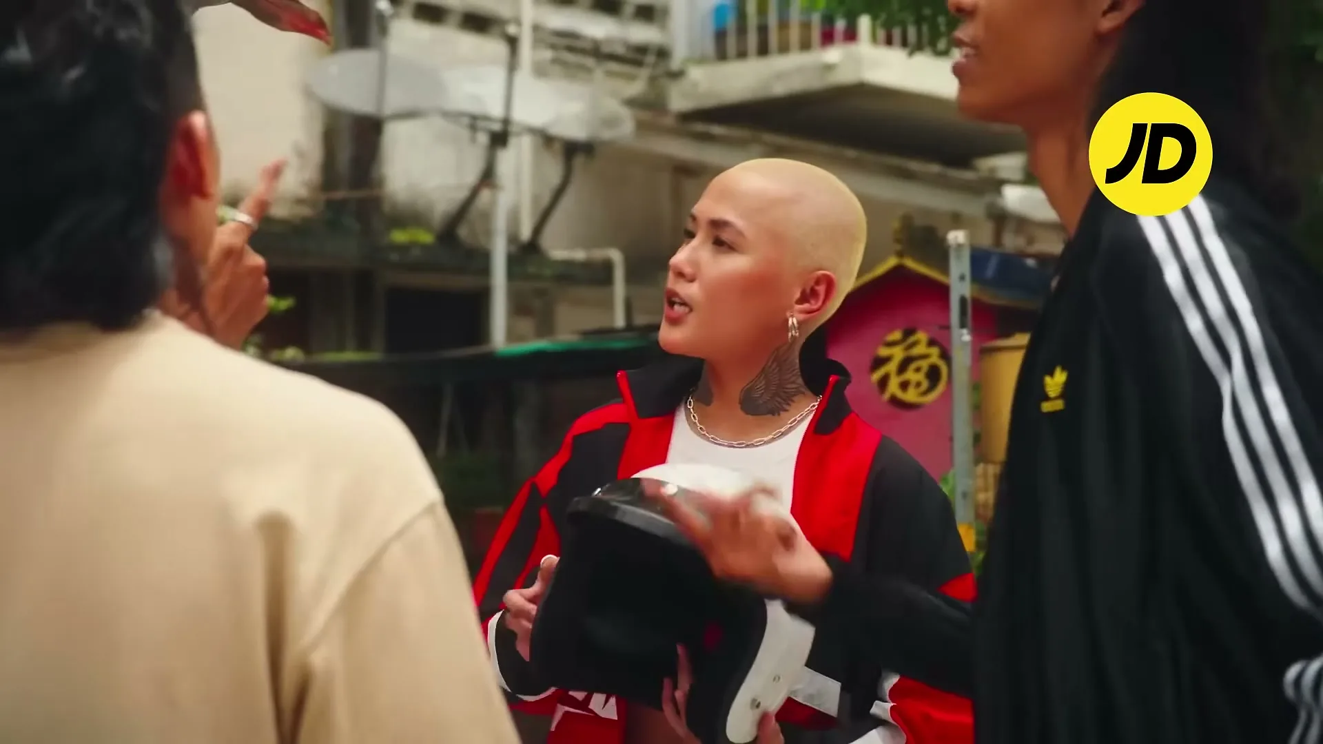 A young woman with a shaved head and tattoos holds a helmet while talking to two other people in an urban setting during the day.