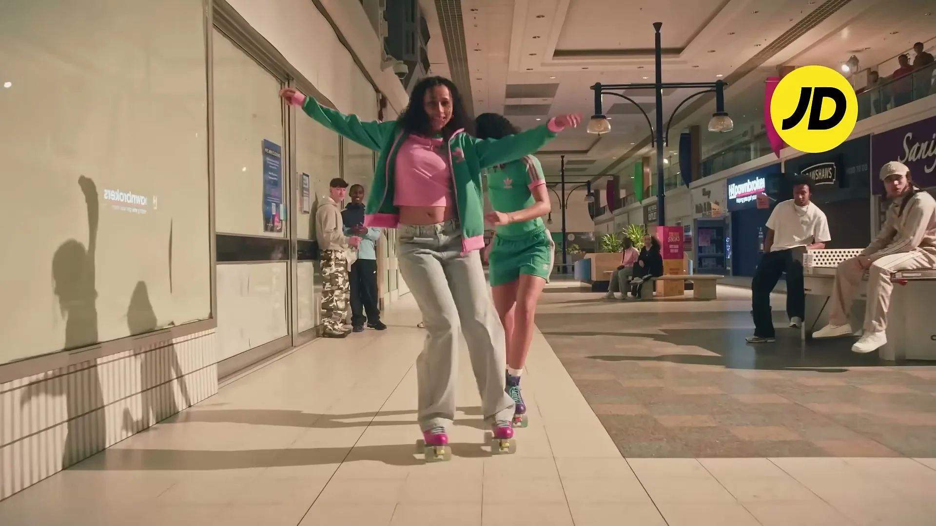 Two young women in athletic wear roller skate through a brightly lit shopping mall, with other people visible in the background.