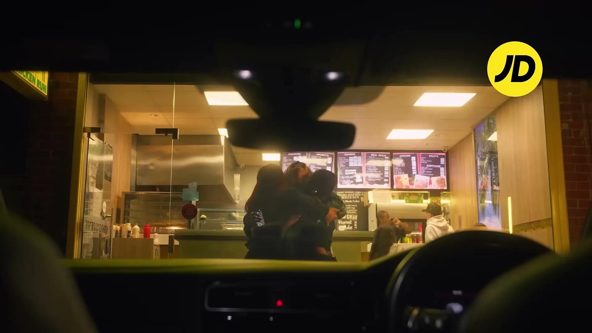 Three young women in casual clothes hug inside a brightly lit fast food restaurant at night, viewed from inside a car.