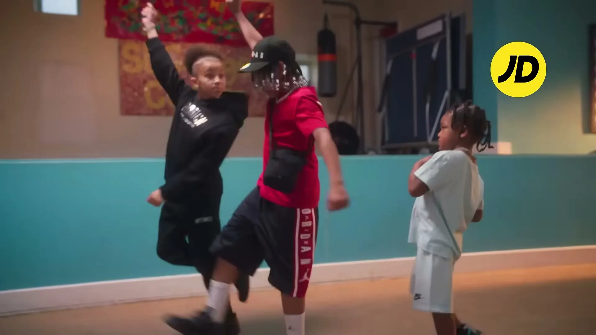 Three children, two boys and one girl, wearing athletic wear, dance energetically in a community center with teal walls.