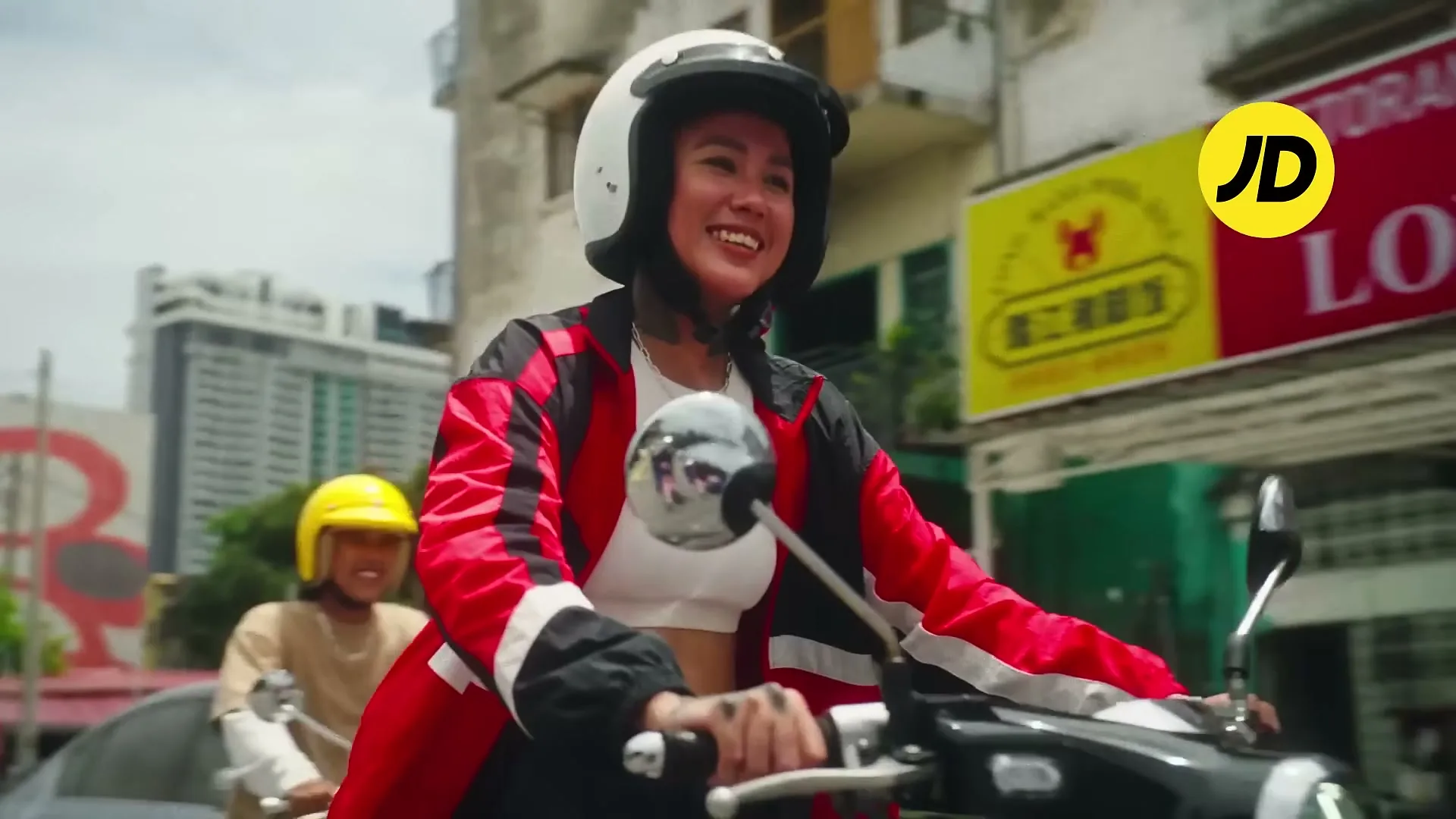 A young woman with tattoos, wearing a helmet and a red and black jacket, smiles while riding a scooter on a city street during the day.