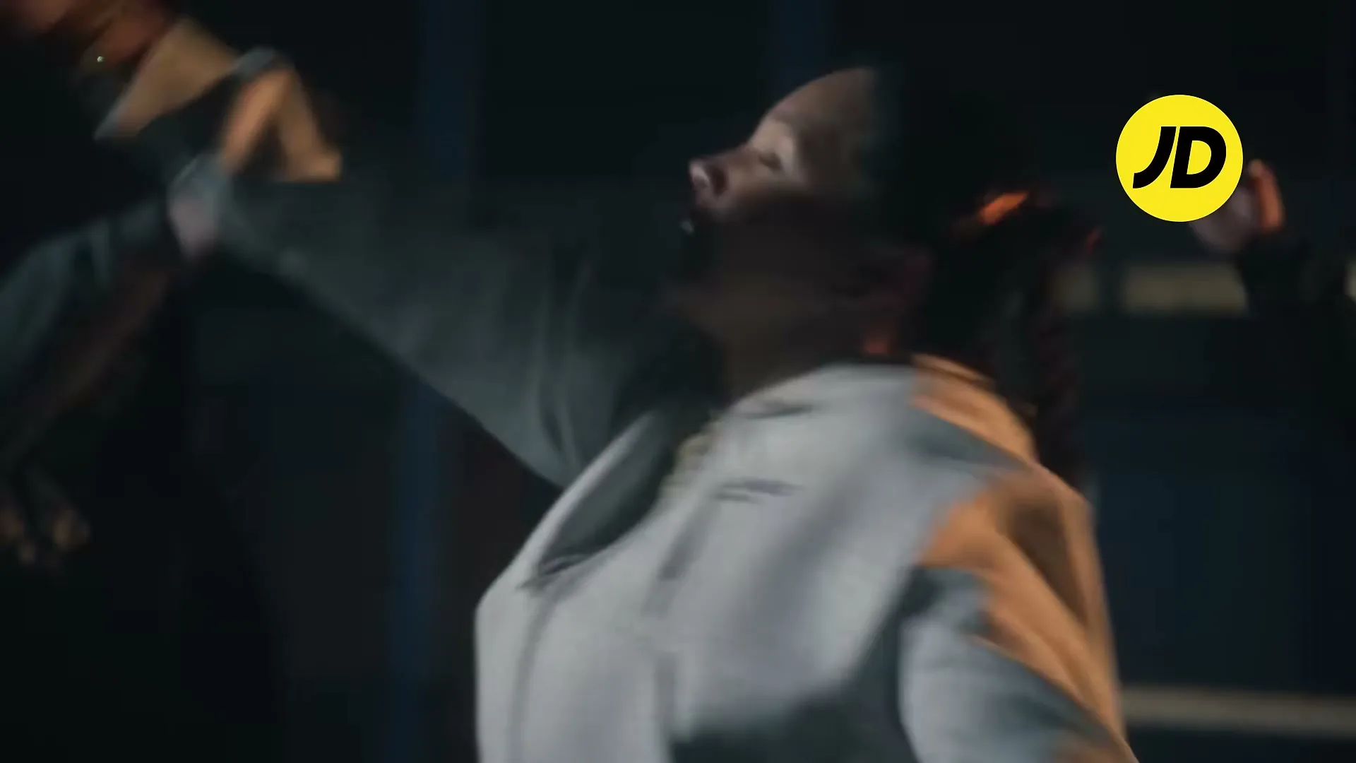 A young woman with braided hair, wearing a grey hoodie, dances energetically with her arms raised in an urban setting at night.