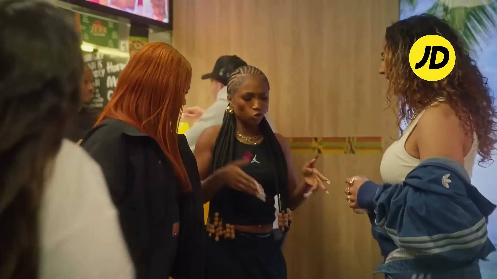 Three women, one with long red hair, one with braids, and one with curly brown hair, talk animatedly in a brightly lit fast-food restaurant.