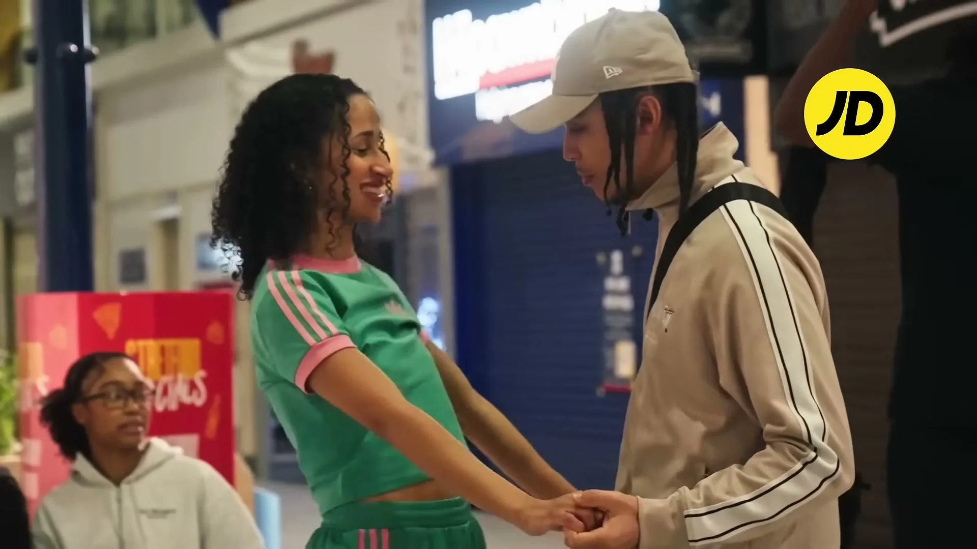 A young woman in a green and pink tracksuit holds hands with a young man in a beige tracksuit and cap, smiling at each other in a brightly lit shopping mall.