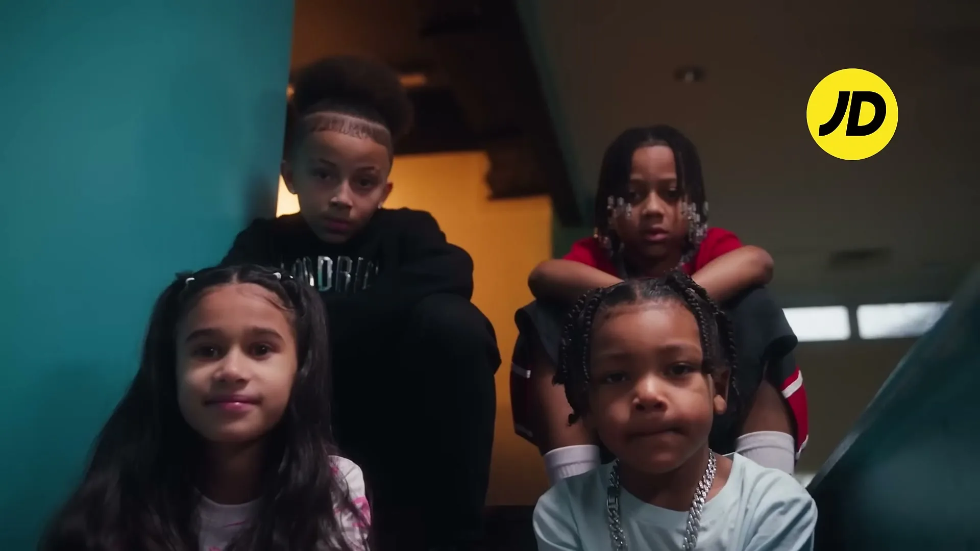 Four children, two boys and two girls, sit on steps looking directly at the camera with serious expressions in an interior setting.