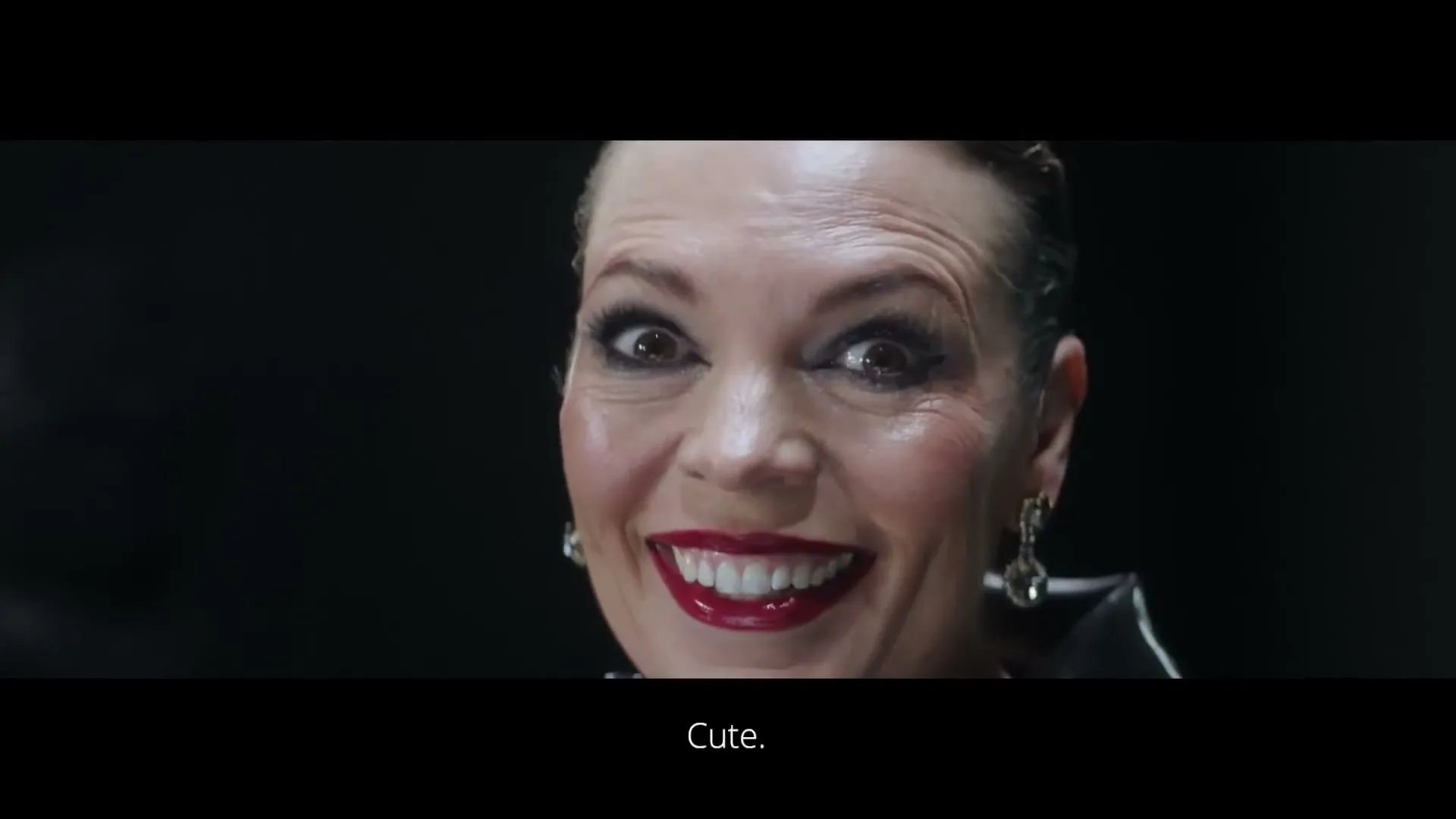 A woman with dark hair and red lipstick smiles mischievously, looking directly at the camera in a dark, indeterminate setting.