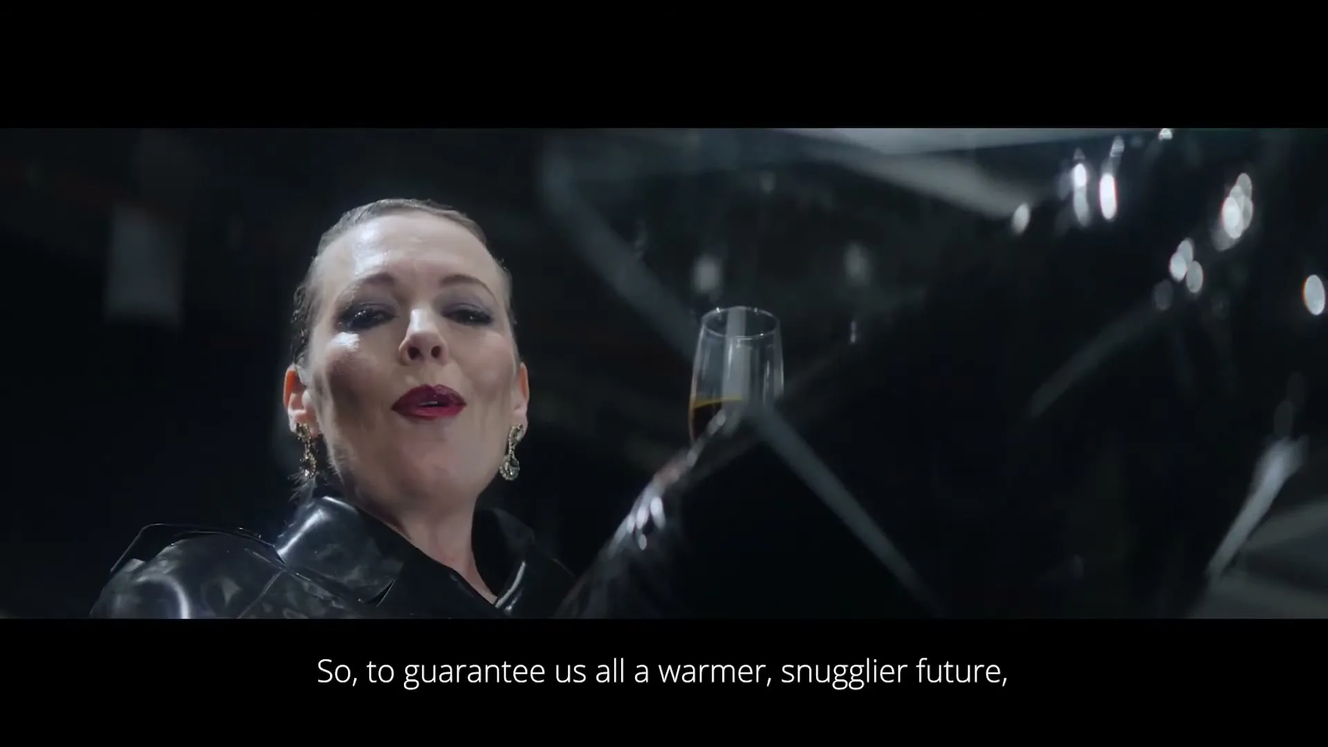 A woman in her 40s, wearing a black latex outfit, holds a glass of dark liquid and smiles wickedly in a dark, industrial setting.