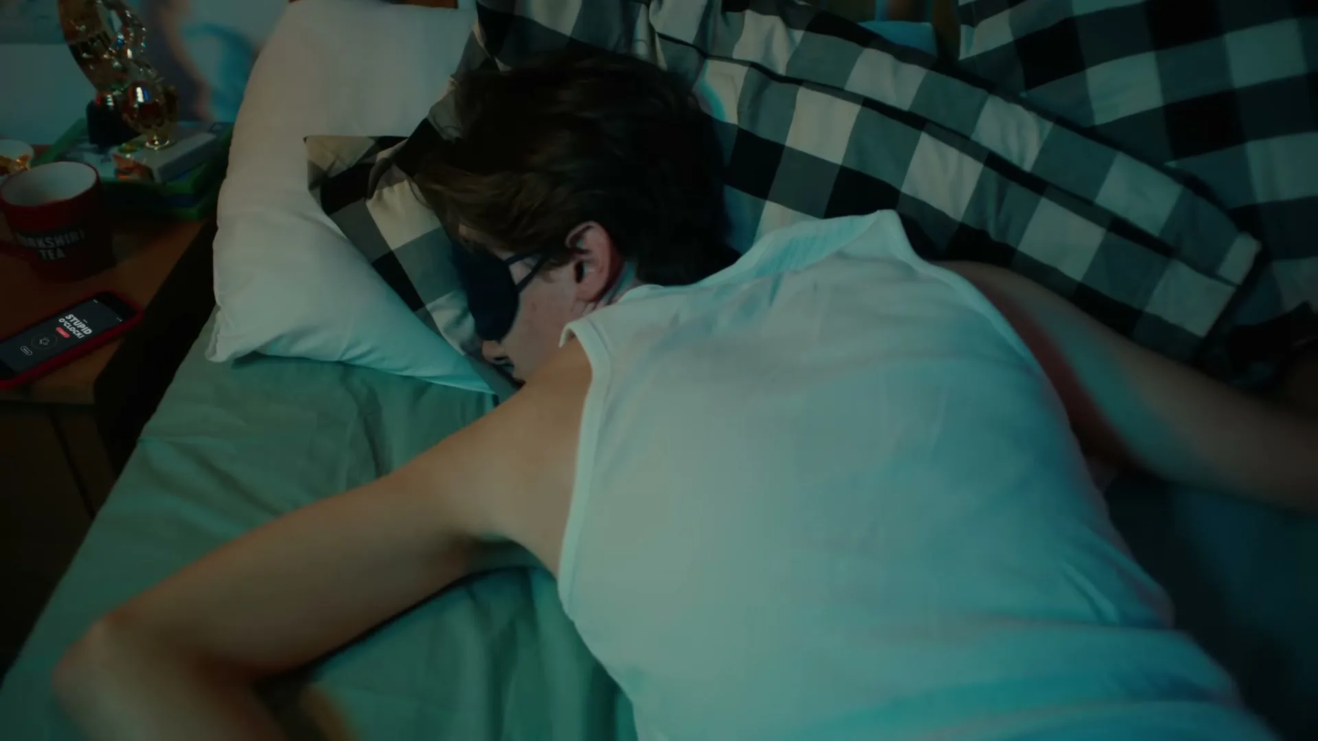 A young man with a sleep mask is sleeping soundly in his bed, with a phone on his nightstand displaying a missed call.