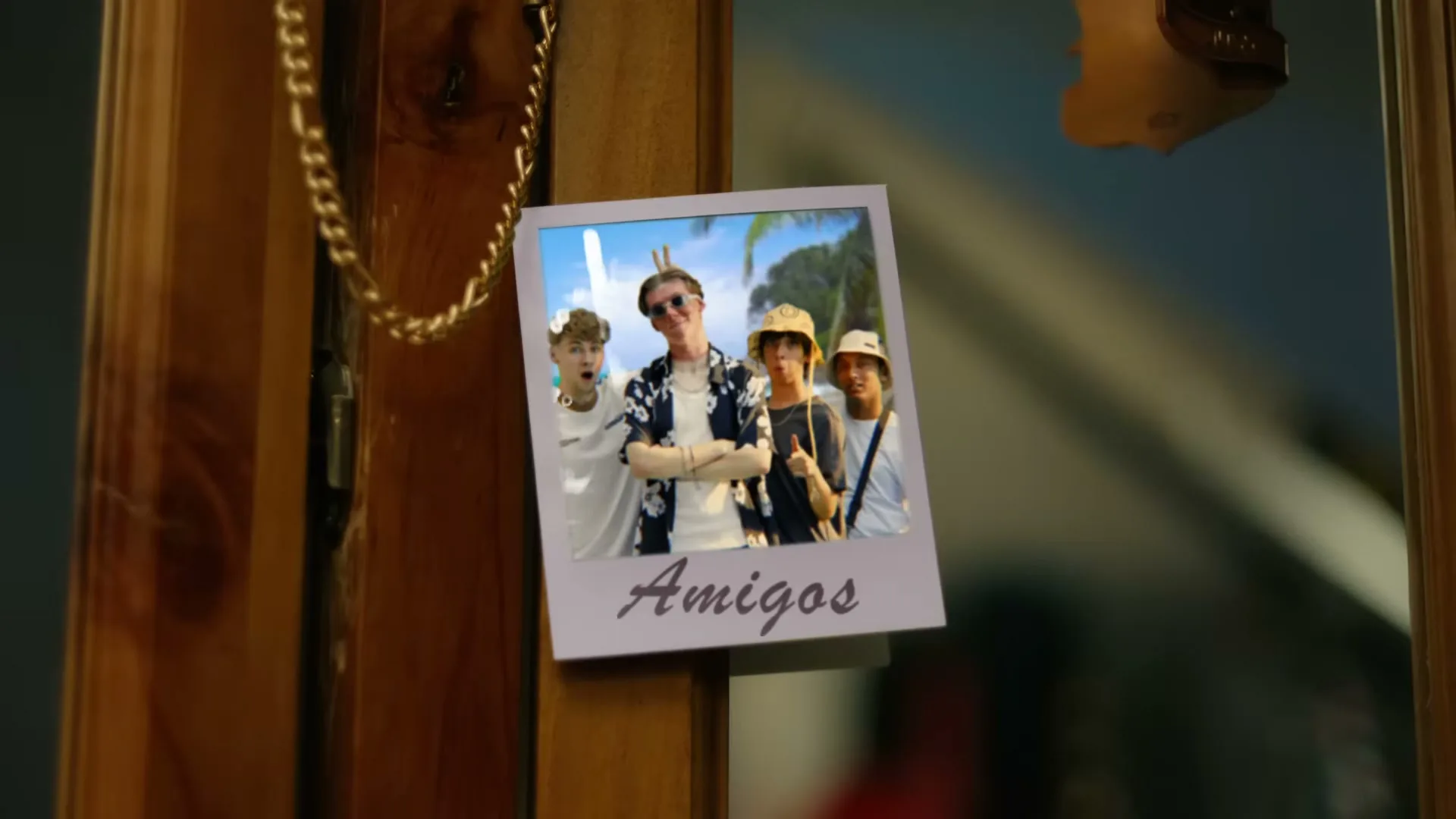 A polaroid photo of four young men on vacation is stuck to a wooden surface with a gold chain hanging nearby.