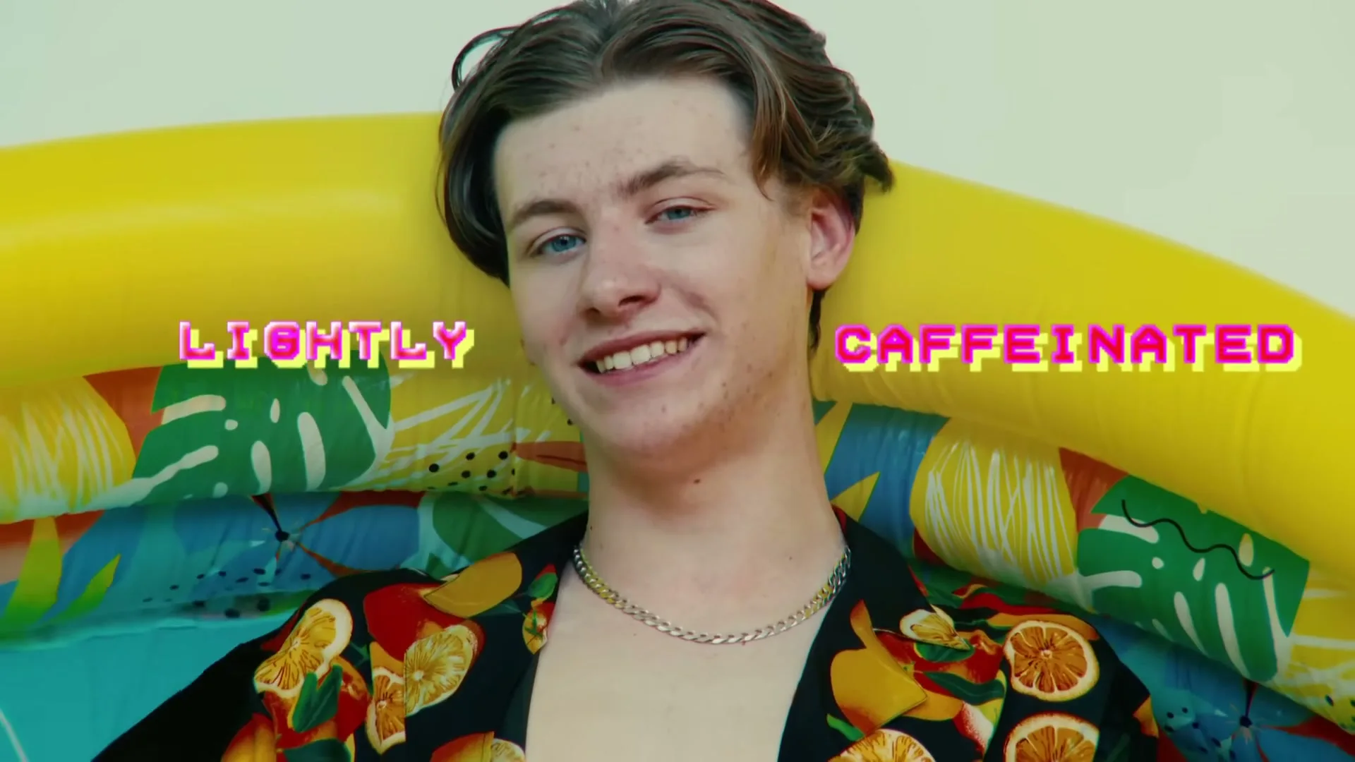 A young man in a Hawaiian shirt and orange shorts sits in a yellow inflatable pool, taking off his sunglasses and smiling at the camera.
