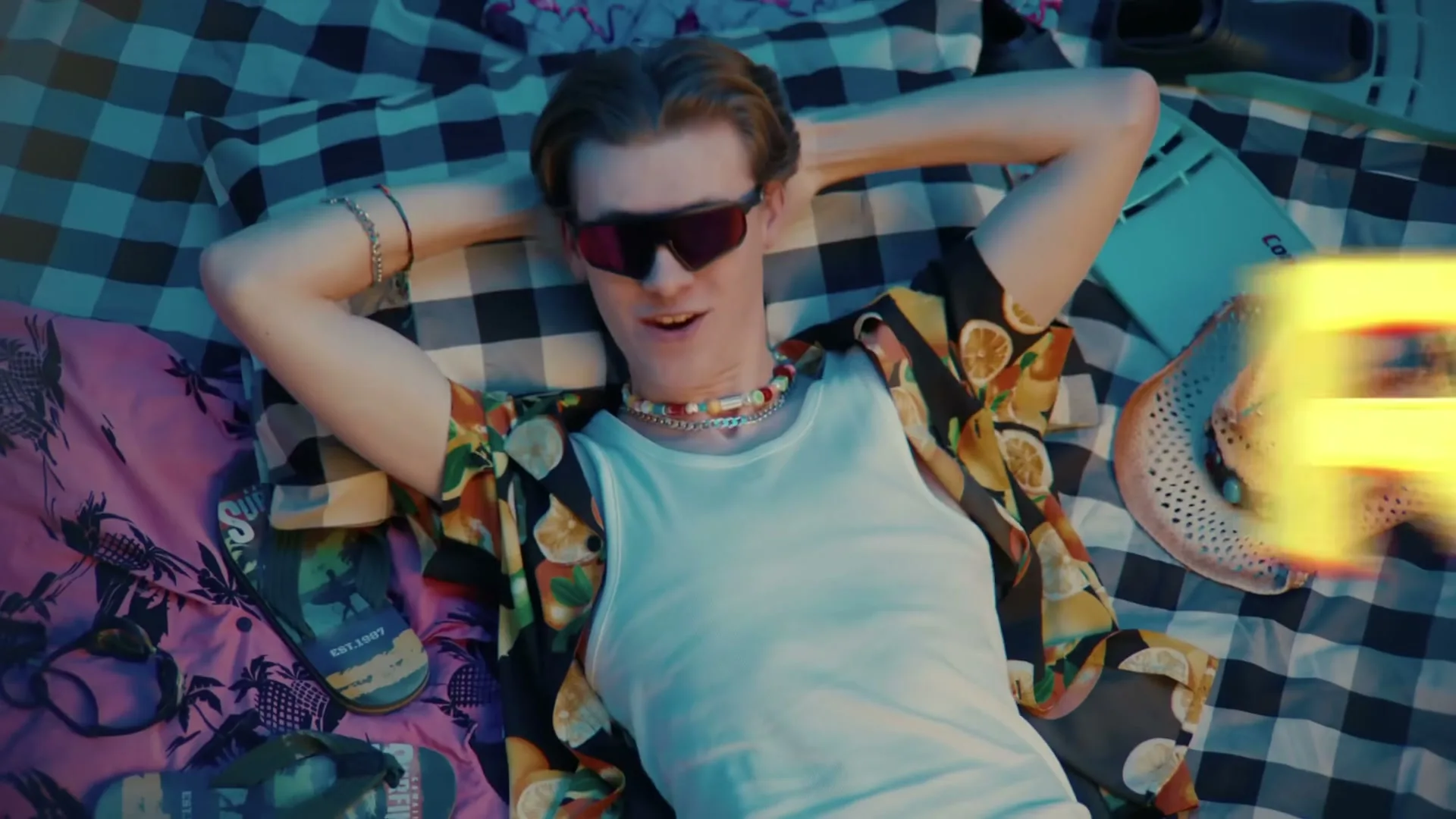 A young man with sunglasses and a colorful shirt lies on a checkered blanket, smiling with text overlayed.