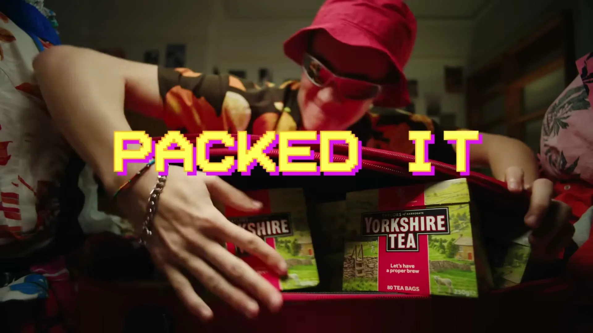 A young man in a red bucket hat and sunglasses packs two boxes of Yorkshire Tea into a red suitcase in a bedroom.
