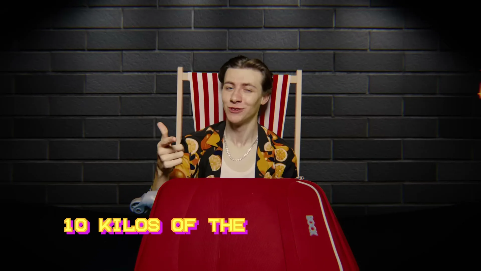 A young man in a Hawaiian shirt sits in a deck chair behind a red suitcase, pointing at the camera with text on screen.