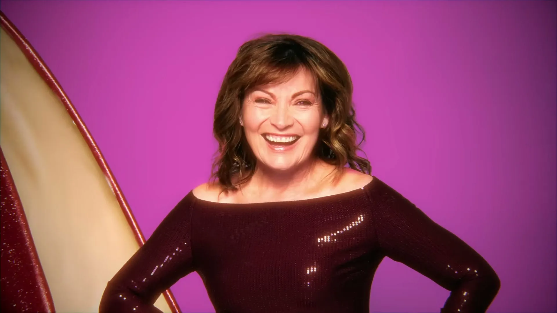A woman in her 60s, Lorraine Kelly, laughs joyfully while wearing a sparkly off-the-shoulder top, standing in a studio with a purple background.