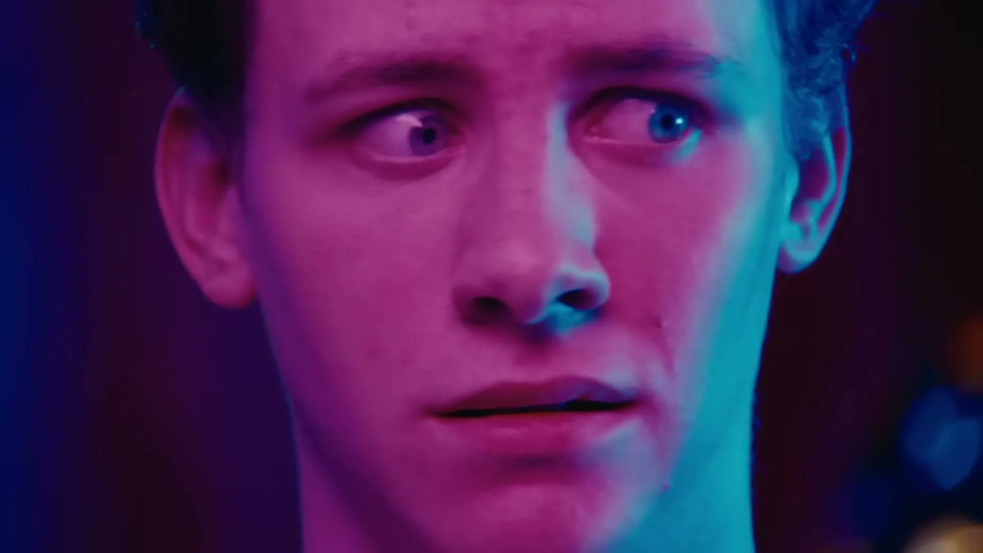 A young man with blue eyes looks around nervously, his face illuminated by pink and blue neon lights in a dark interior.