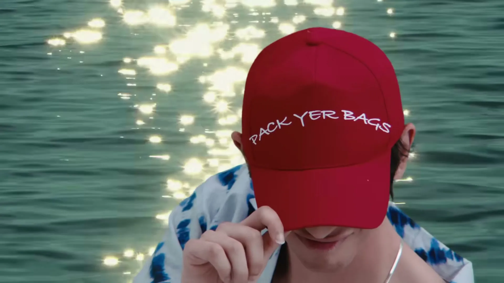 A young man in a red cap with "PACK YER BAGS" written on it, and a Hawaiian shirt, looks down at sparkling water.
