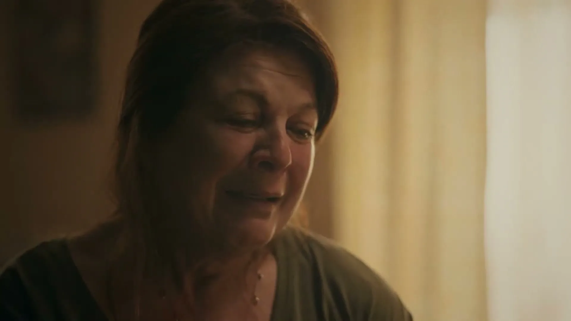 A woman in her 60s with short brown hair and a green shirt looks down and smiles, then her expression changes to sadness, in a dimly lit room.