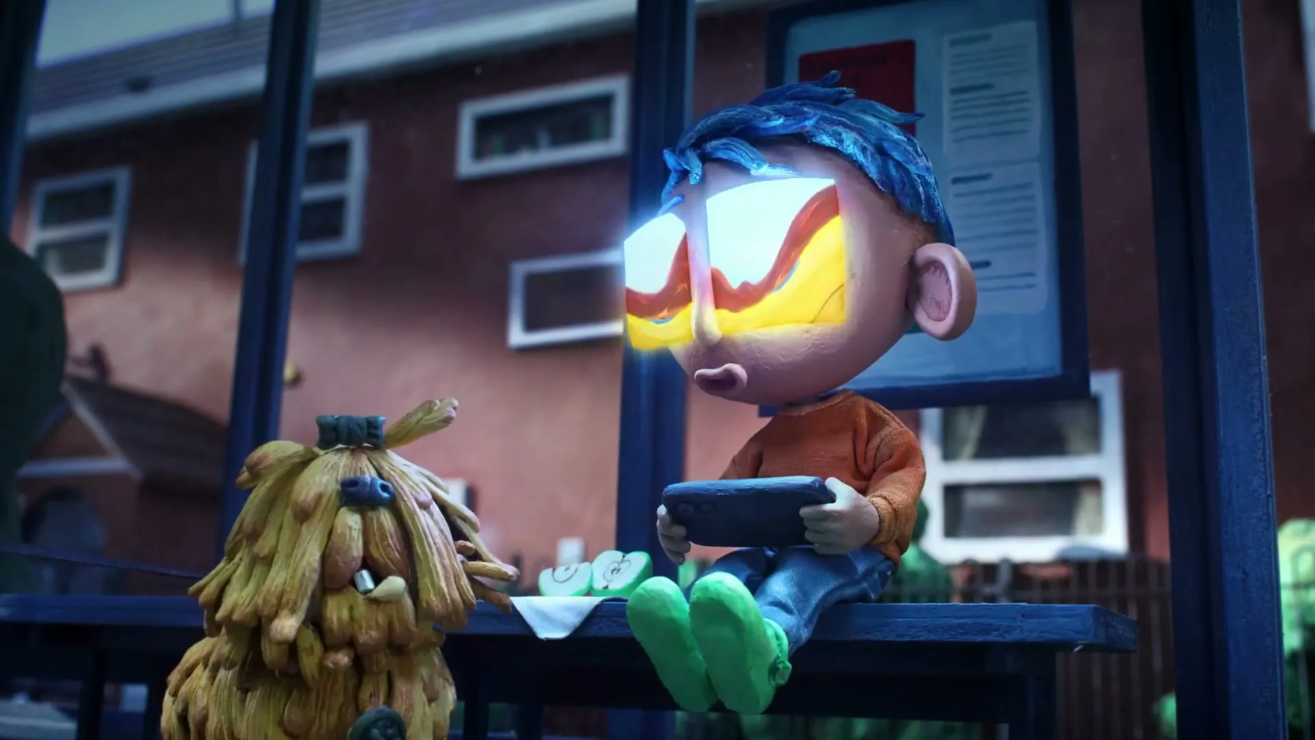A claymation boy with glowing eyes sits next to a shaggy dog, holding a game controller, in a window at night.