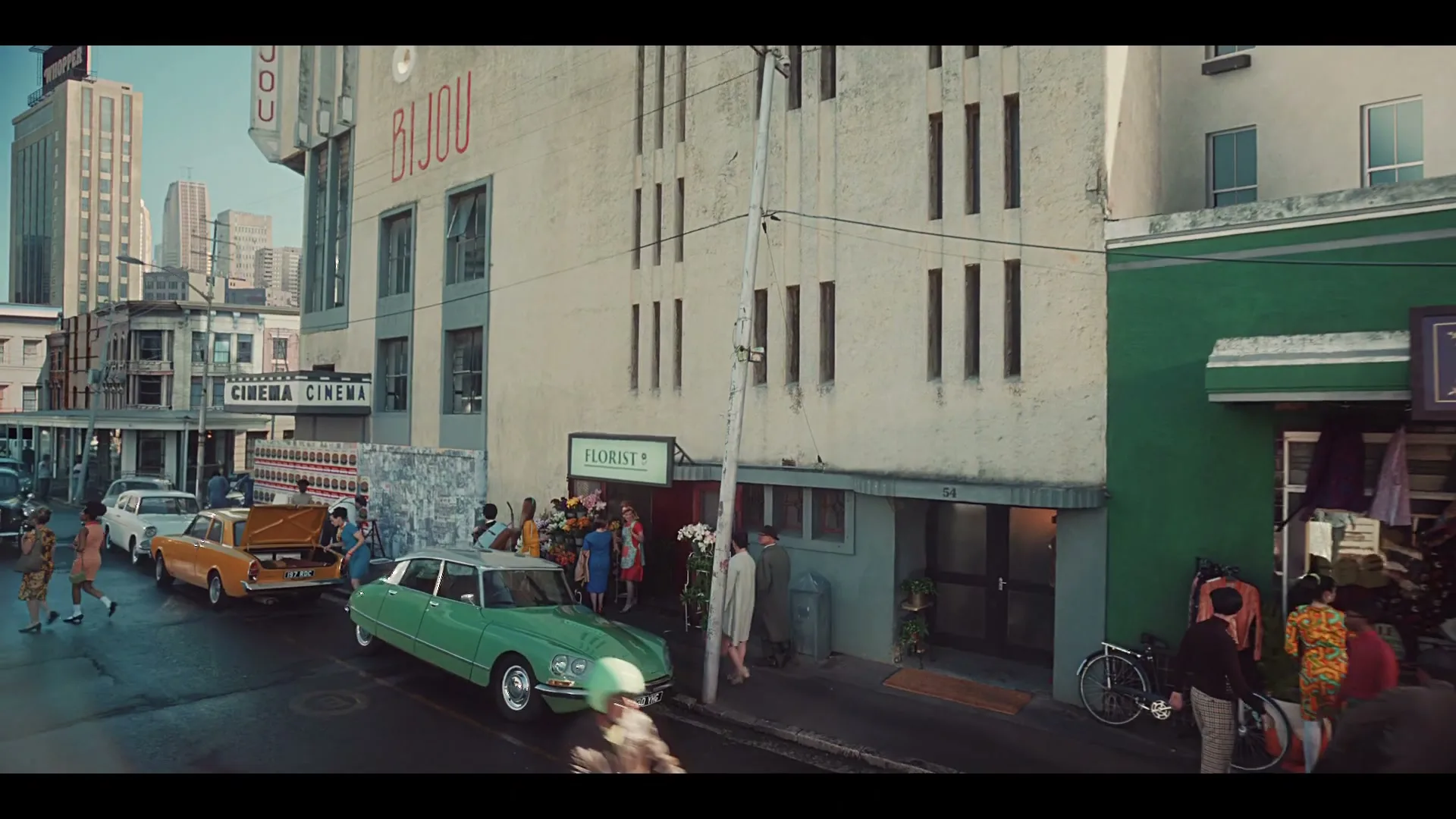 A bustling street scene in a retro city, with people walking, a green car, and a scooter, in front of a florist and a cinema.