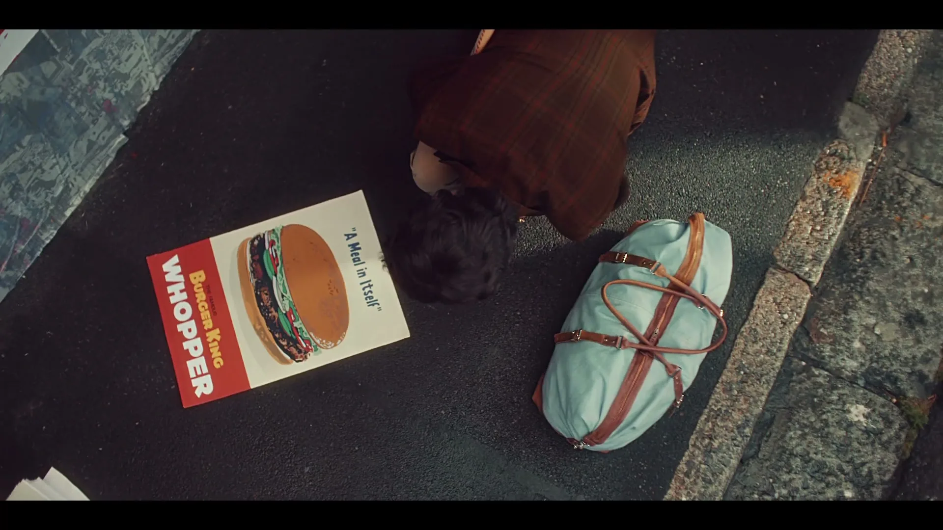 A man in a brown suit kneels on the ground next to a duffel bag and a Burger King Whopper sign on a sidewalk.