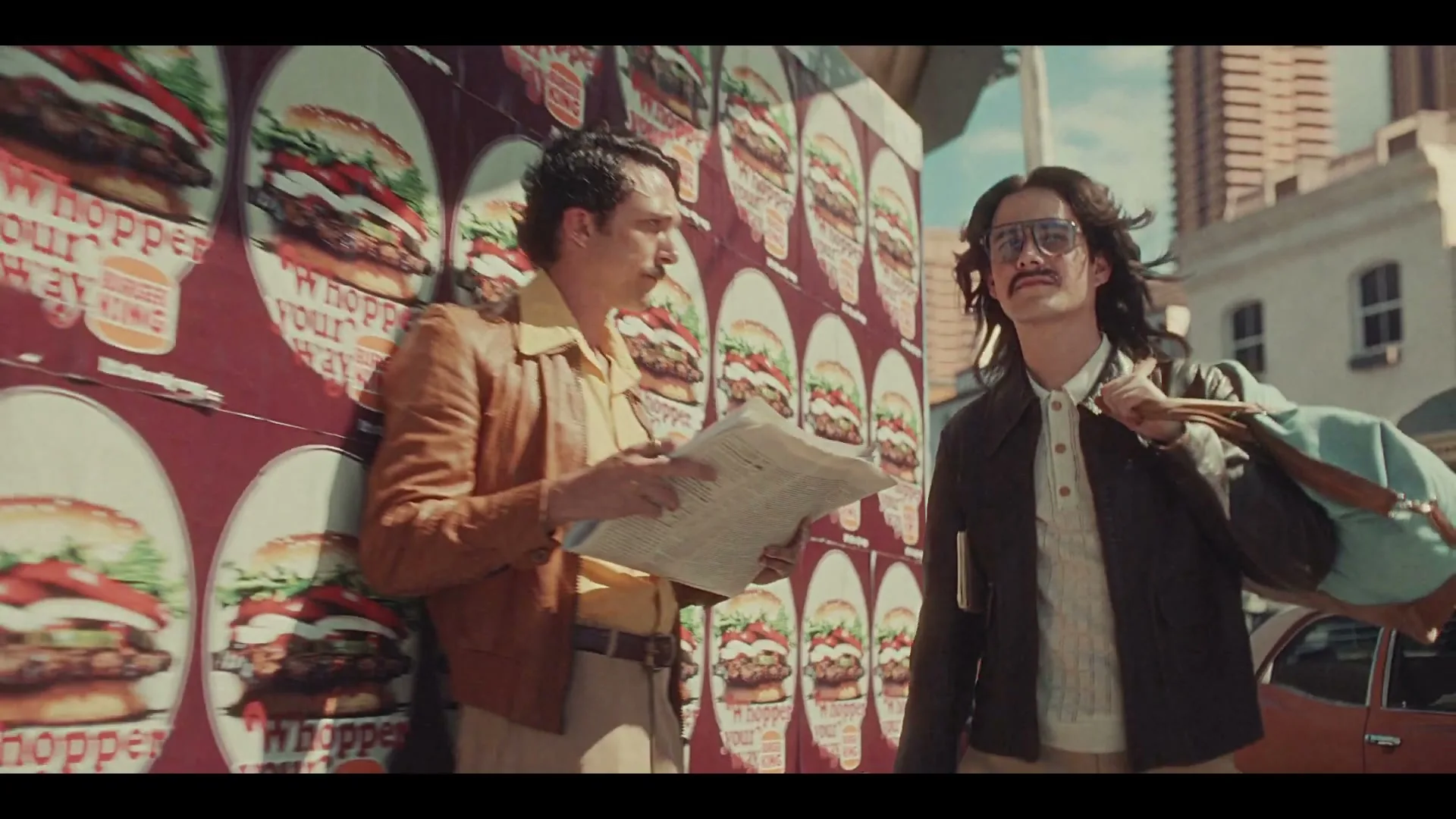 Two men with 70s hairstyles and clothing stand next to a wall covered in Burger King Whopper posters on a city street.