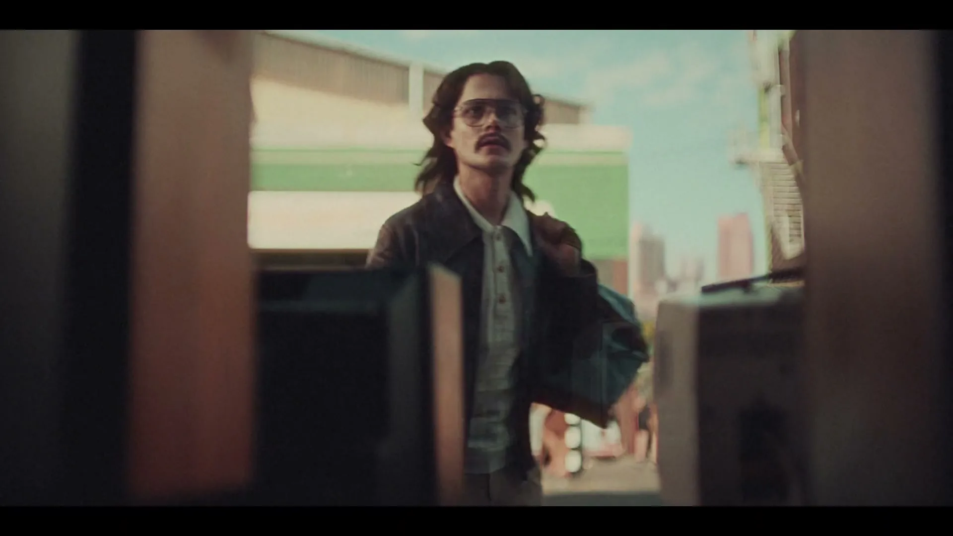 A man with long hair and a mustache, wearing a jacket and glasses, walks towards the camera on a sunny street, adjusting his collar.